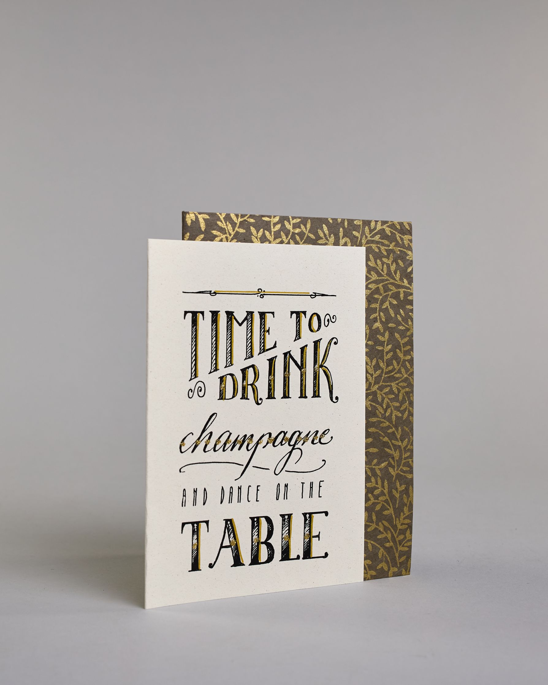 Karte in Englisch mit der Aufschrift „Time to drink champagne and dance on the table“ in dekorativer Handlettering-Schrift auf hellem Hintergrund. Die Wörter sind in Schwarz gestaltet, teils mit goldfarbenen und glitzernden Details hervorgehoben. Das "TIME TO DRINK" und "TABLE" ist in kreativer gerader Kalligrafieaufschrift gestaltet, das "Champagne" in geschwungener und das "AND DANCE ON THE" in dünner gerader Schrift. Dazu ein grün-goldenes handgeschöpftes Kuvert. #3