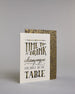 Karte in Englisch mit der Aufschrift „Time to drink champagne and dance on the table“ in dekorativer Handlettering-Schrift auf hellem Hintergrund. Die Wörter sind in Schwarz gestaltet, teils mit goldfarbenen und glitzernden Details hervorgehoben. Das "TIME TO DRINK" und "TABLE" ist in kreativer gerader Kalligrafieaufschrift gestaltet, das "Champagne" in geschwungener und das "AND DANCE ON THE" in dünner gerader Schrift. Dazu ein grün-goldenes handgeschöpftes Kuvert. #3