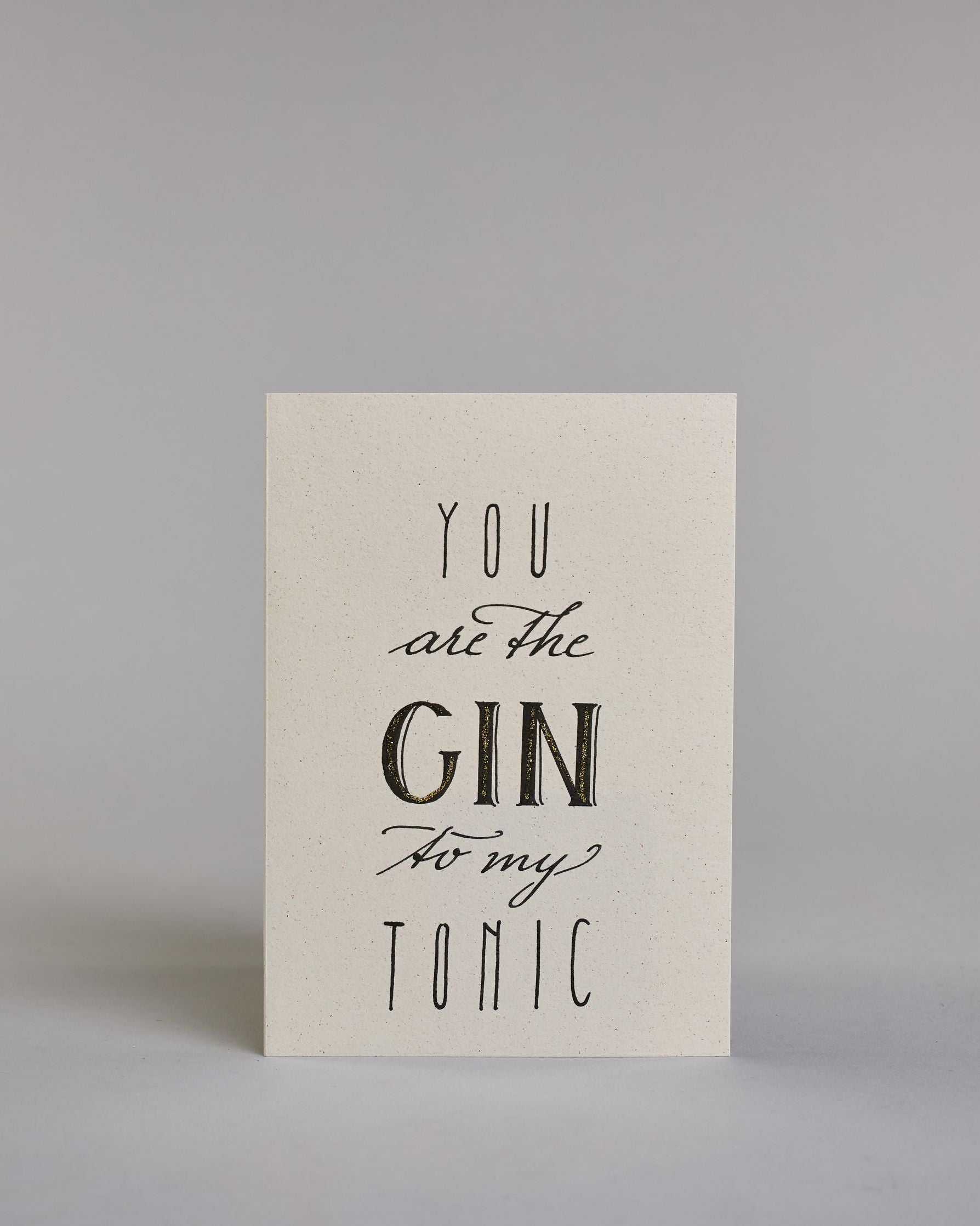 Karte in Englisch mit der Aufschrift „You are the Gin to my Tonic“ in schwarzer Handlettering-Schrift, wobei das Wort „GIN“ mit goldfarbenen Details hervorgehoben ist. Die Zeilen setzten sich wiefolgt zusammen: "YOU" in dünner gerader Schrift, darunte "are the" in geschwungener Schrift, darunter "GIN" in gerader Schrift, darunter "to my" in geschwungener,darunter "TONIC" wieder in dünner gerader Schrift auf hellem Hintergrund. Dazu ein weiß-goldenes Kuvert. #1
