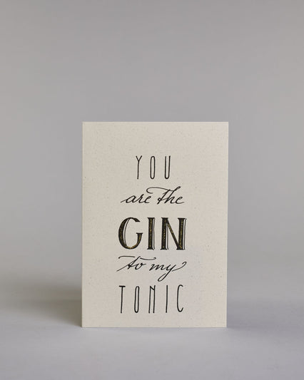 Karte in Englisch mit der Aufschrift „You are the Gin to my Tonic“ in schwarzer Handlettering-Schrift, wobei das Wort „GIN“ mit goldfarbenen Details hervorgehoben ist. Die Zeilen setzten sich wiefolgt zusammen: "YOU" in dünner gerader Schrift, darunte "are the" in geschwungener Schrift, darunter "GIN" in gerader Schrift, darunter "to my" in geschwungener,darunter "TONIC" wieder in dünner gerader Schrift auf hellem Hintergrund. Dazu ein weiß-goldenes Kuvert. #1