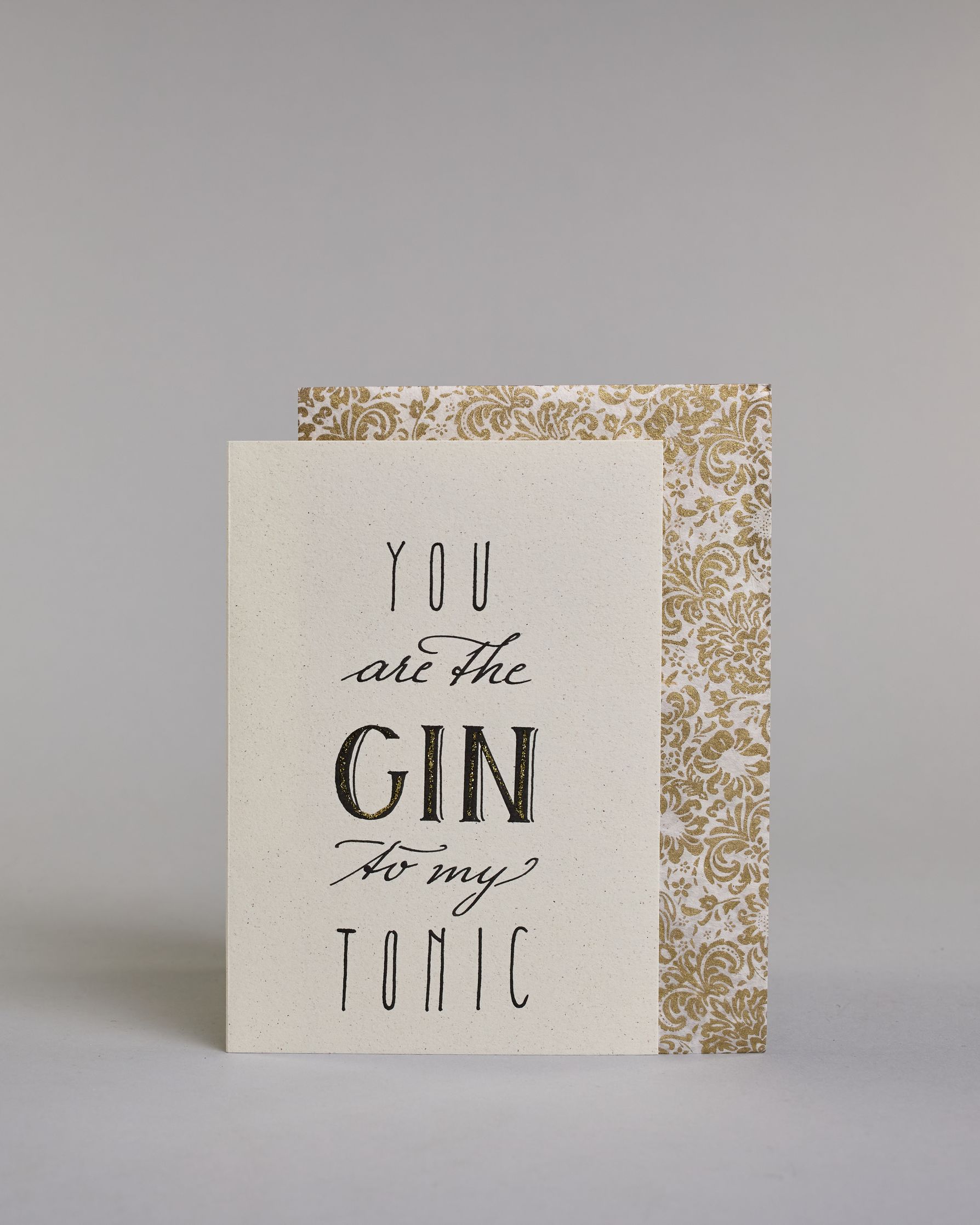 Karte in Englisch mit der Aufschrift „You are the Gin to my Tonic“ in schwarzer Handlettering-Schrift, wobei das Wort „GIN“ mit goldfarbenen Details hervorgehoben ist. Die Zeilen setzten sich wiefolgt zusammen: "YOU" in dünner gerader Schrift, darunte "are the" in geschwungener Schrift, darunter "GIN" in gerader Schrift, darunter "to my" in geschwungener,darunter "TONIC" wieder in dünner gerader Schrift auf hellem Hintergrund. Dazu ein weiß-goldenes Kuvert. #2