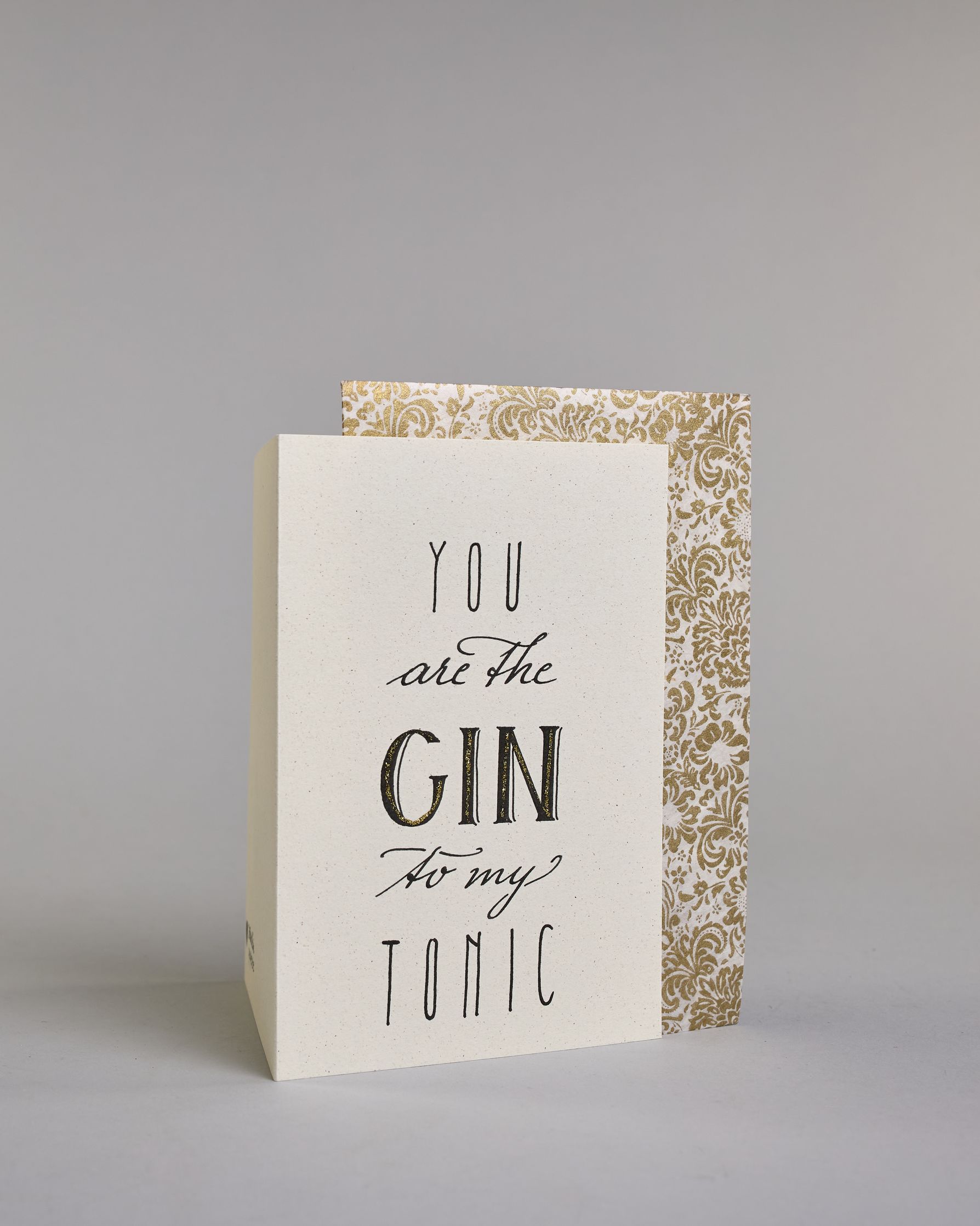 Karte in Englisch mit der Aufschrift „You are the Gin to my Tonic“ in schwarzer Handlettering-Schrift, wobei das Wort „GIN“ mit goldfarbenen Details hervorgehoben ist. Die Zeilen setzten sich wiefolgt zusammen: "YOU" in dünner gerader Schrift, darunte "are the" in geschwungener Schrift, darunter "GIN" in gerader Schrift, darunter "to my" in geschwungener,darunter "TONIC" wieder in dünner gerader Schrift auf hellem Hintergrund. Dazu ein weiß-goldenes Kuvert. #3