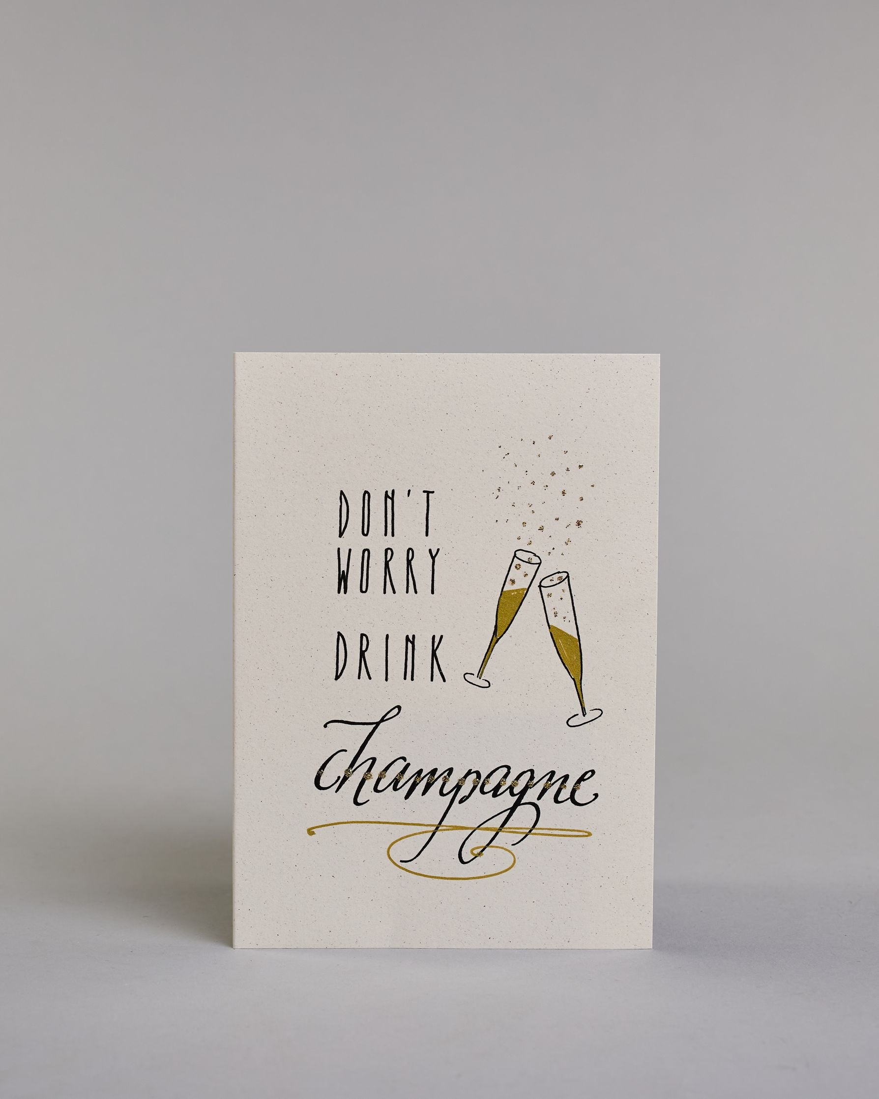 Karte in Englisch mit der Aufschrift „Don’t worry drink champagne“ in schwarzer Handlettering-Schrift. Daneben sind zwei angestoßene goldene verzierte Champagnergläser mit aufsteigenden Bläschen illustriert. Das "DON'T WORRY DRINK" ist auf drei untereinanderliegenden Zeilen in gerader und das "champagne" darunter in geschwungener Schrift auf hellem Hintergrund dargestellt. Dazu ein weiß-goldenes Kuvert. #1