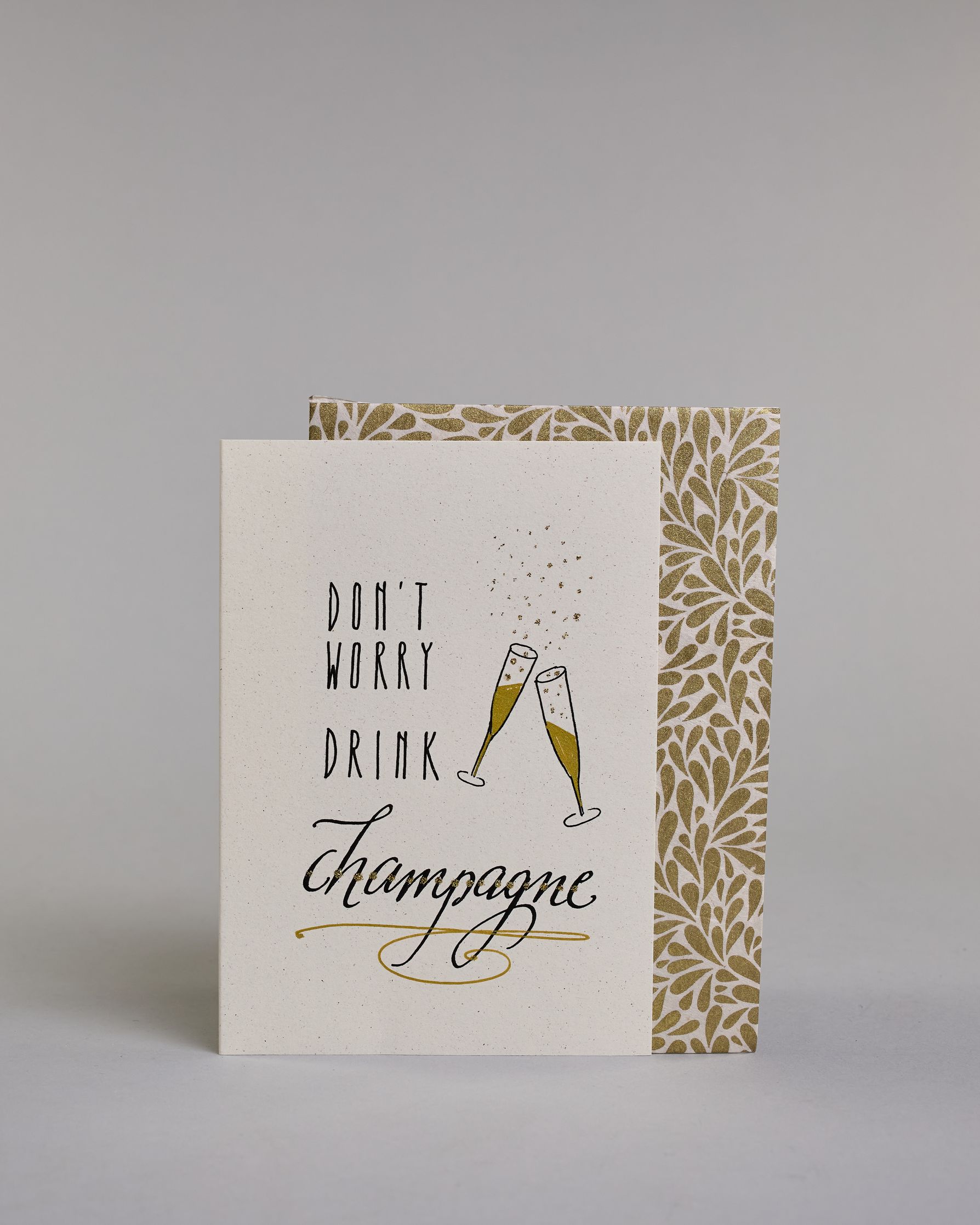 Karte in Englisch mit der Aufschrift „Don’t worry drink champagne“ in schwarzer Handlettering-Schrift. Daneben sind zwei angestoßene goldene verzierte Champagnergläser mit aufsteigenden Bläschen illustriert. Das "DON'T WORRY DRINK" ist auf drei untereinanderliegenden Zeilen in gerader und das "champagne" darunter in geschwungener Schrift auf hellem Hintergrund dargestellt. Dazu ein weiß-goldenes Kuvert. #2
