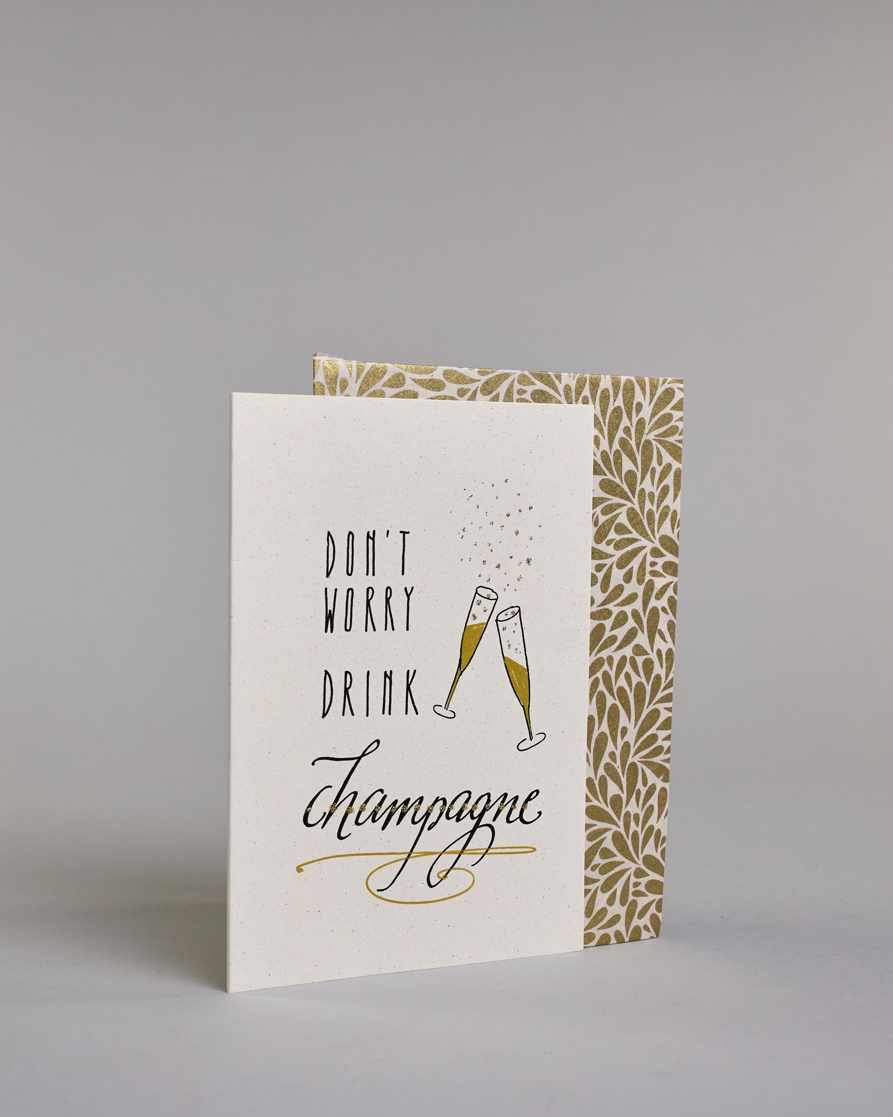 Karte in Englisch mit der Aufschrift „Don’t worry drink champagne“ in schwarzer Handlettering-Schrift. Daneben sind zwei angestoßene goldene verzierte Champagnergläser mit aufsteigenden Bläschen illustriert. Das "DON'T WORRY DRINK" ist auf drei untereinanderliegenden Zeilen in gerader und das "champagne" darunter in geschwungener Schrift auf hellem Hintergrund dargestellt. Dazu ein weiß-goldenes Kuvert. #3
