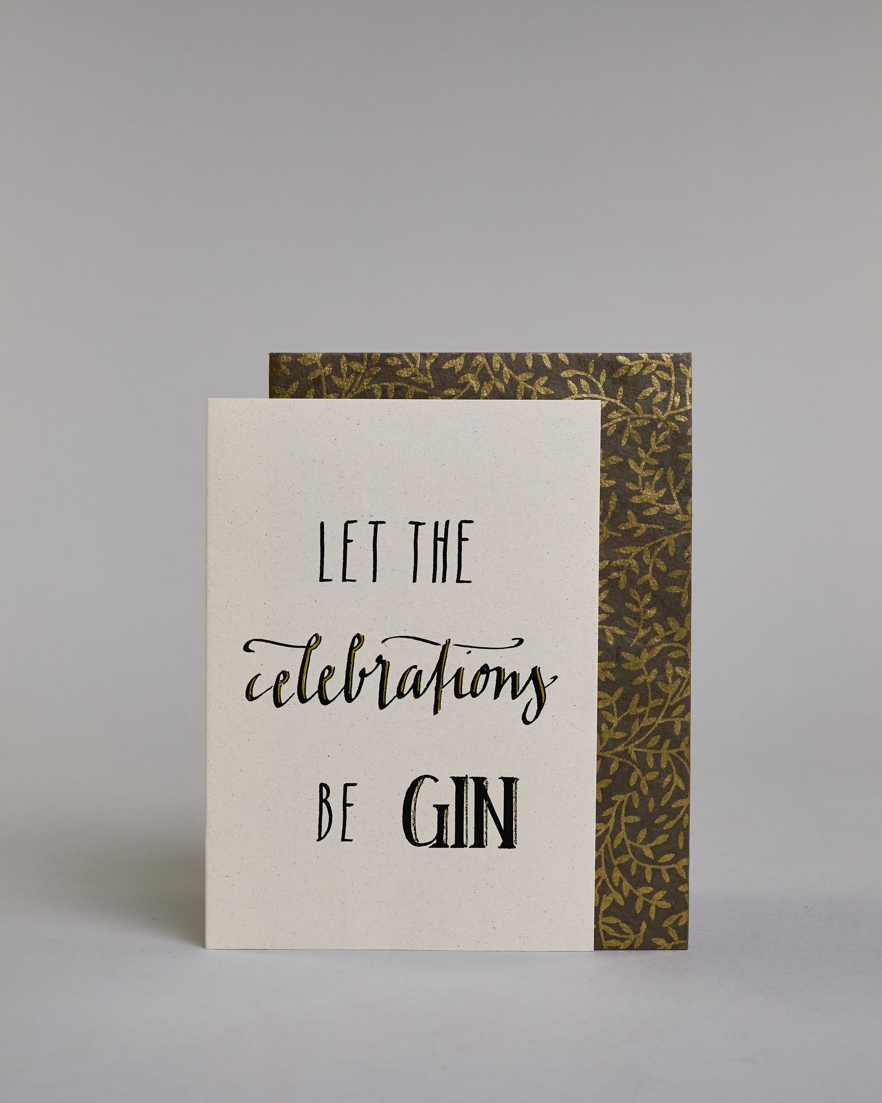 Karte in Englisch mit der Aufschrift „Let the celebrations be gin“ in schwarzer Handlettering-Schrift, wobei die Wörter „celebrations“ und „GIN“ mit goldfarbenen Akzenten hervorgehoben sind. Das "celebrations" steht in der mittleren der drei Zeilen und ist in geschwungener Kalligrafieaufschrift gemacht, während die Zeile darüber und darunter mit dem "LET THE" und "BE GIN"  in gerade Schrift auf hellem Hintergrund sind. Dazu ein grün-goldenes handgeschöpftes Kuvert. #2