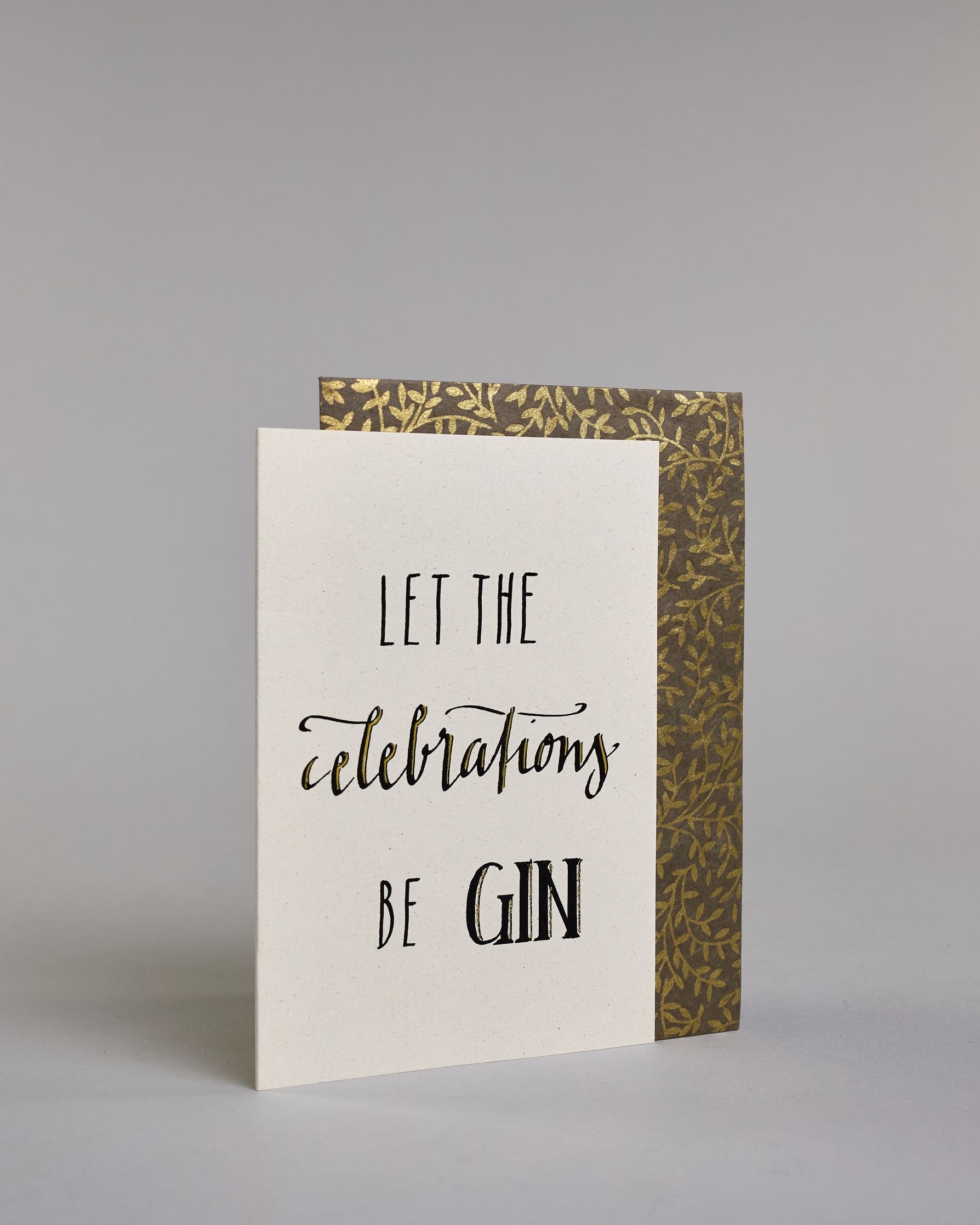 Karte in Englisch mit der Aufschrift „Let the celebrations be gin“ in schwarzer Handlettering-Schrift, wobei die Wörter „celebrations“ und „GIN“ mit goldfarbenen Akzenten hervorgehoben sind. Das "celebrations" steht in der mittleren der drei Zeilen und ist in geschwungener Kalligrafieaufschrift gemacht, während die Zeile darüber und darunter mit dem "LET THE" und "BE GIN"  in gerade Schrift auf hellem Hintergrund sind. Dazu ein grün-goldenes handgeschöpftes Kuvert. #3