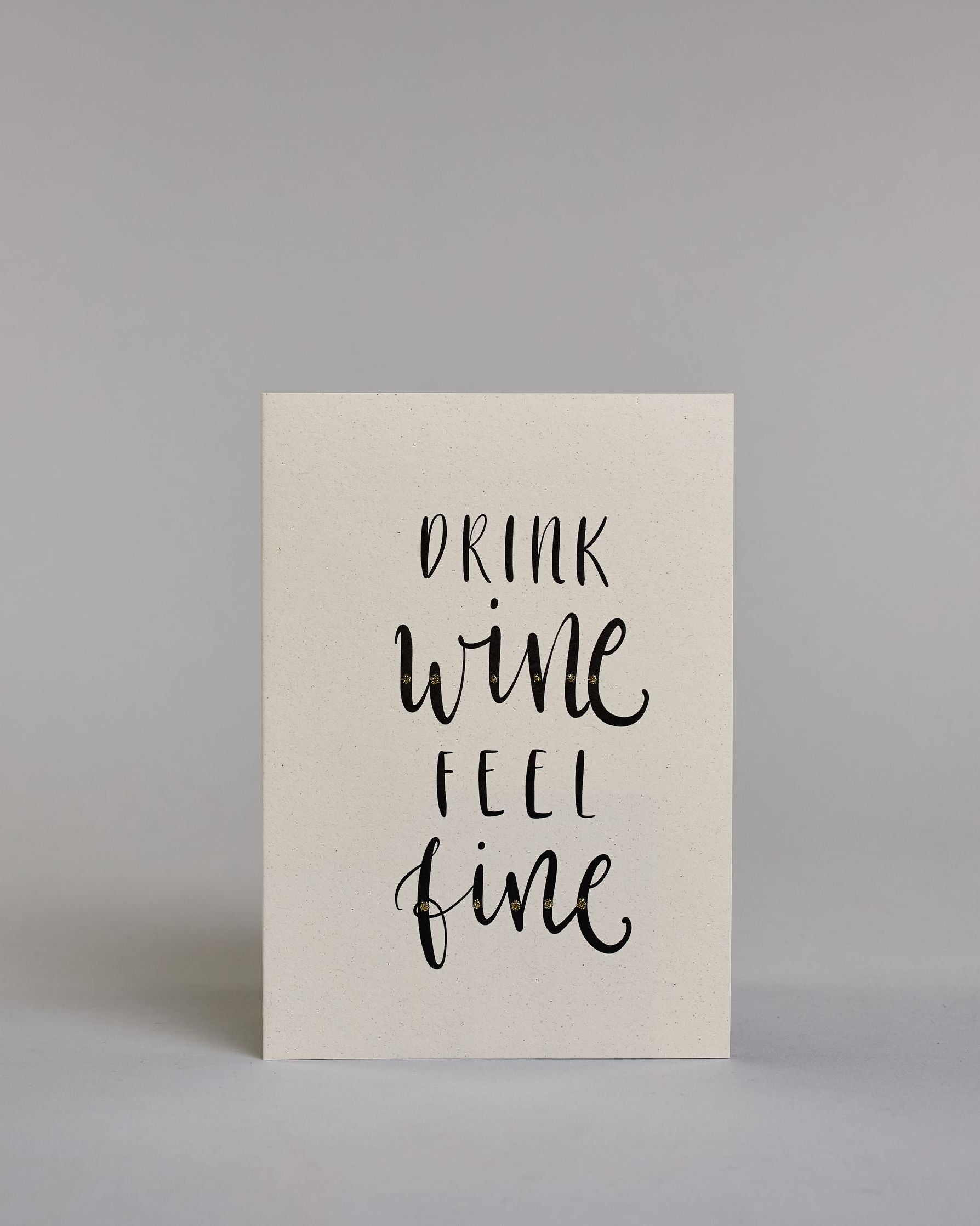 Karte in Englisch mit der Aufschrift „Drink wine feel fine“ in schwarzer Handlettering-Schrift, teils mit kleinen goldfarbenen Akzenten auf vier Zeilen mit jeweils immer einem Wort aufgeteilt und auf hellem Hintergrund. Dazu ein grün-goldenes handgeschöpftes Kuvert. #1