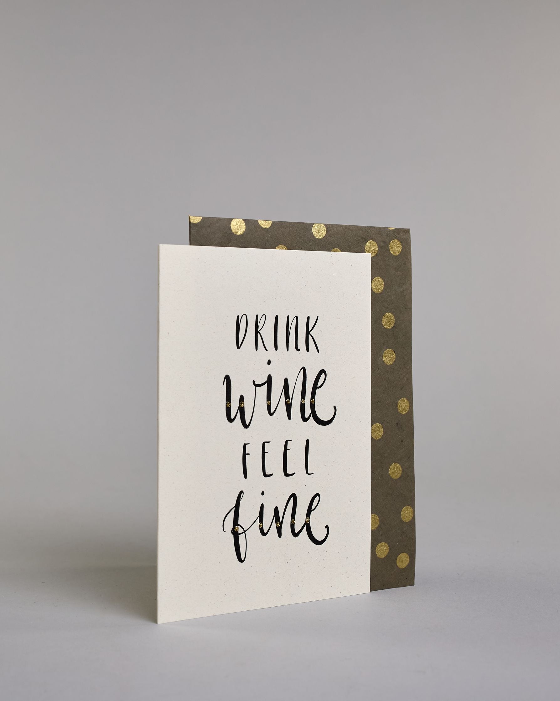 Karte in Englisch mit der Aufschrift „Drink wine feel fine“ in schwarzer Handlettering-Schrift, teils mit kleinen goldfarbenen Akzenten auf vier Zeilen mit jeweils immer einem Wort aufgeteilt und auf hellem Hintergrund. Dazu ein grün-goldenes handgeschöpftes Kuvert. #3