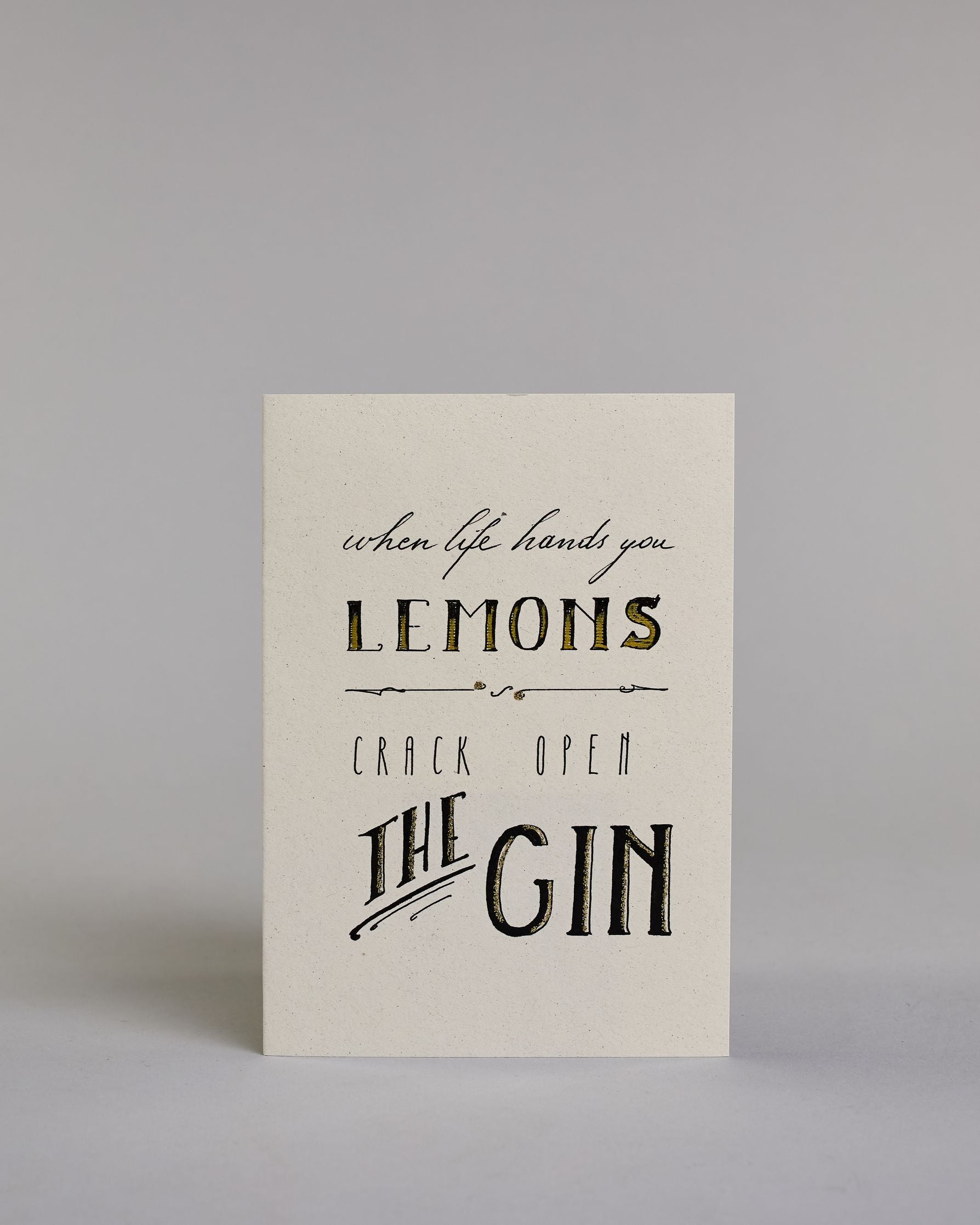 Karte in Englisch mit der Aufschrift „When life hands you lemons crack open the gin“ in schwarzer Handlettering-Schrift, wobei die Wörter „LEMONS“ und „THE GIN“ mit goldfarbenen Schattierungen hervorgehoben sind. Die oberen zwei Zeilen, "when life hands you" und "LEMONS" sind durch eine horizontale kalligrafische Linie von den unteren beiden Zeilen "CRACK OPEN" und "THE GIN" getrennt und auf hellem Hintergrund dargestellt. Dazu ein grün-goldenes handgeschöpftes Kuvert. #1
