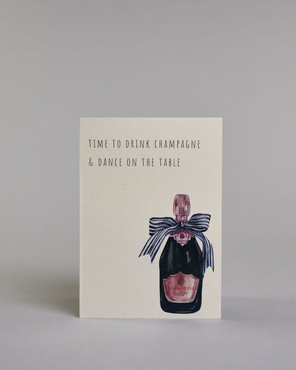 Karte in Englisch mit der Aufschrift „Time to drink champagne & dance on the table“ in schlichter schwarzer Kalligrafieaufschrift. Rechts unten auf der Karte ist eine illustrierte Champagnerflasche mit rosa Etikett und Verschluss zu sehen, geschmückt mit einer großen dunkelblauen Schleife. Dazu ein rosa-weißes handgeschöpftes Kuvert. #1