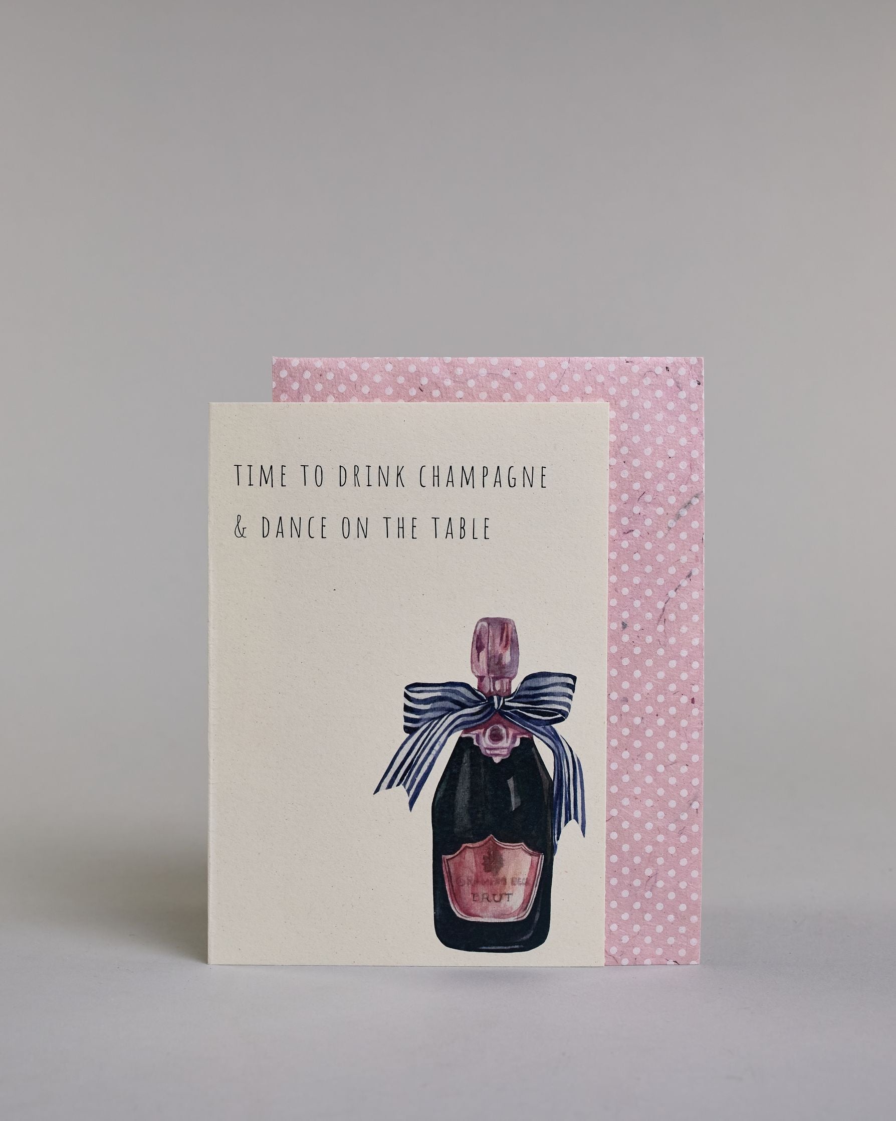 Karte in Englisch mit der Aufschrift „Time to drink champagne & dance on the table“ in schlichter schwarzer Kalligrafieaufschrift. Rechts unten auf der Karte ist eine illustrierte Champagnerflasche mit rosa Etikett und Verschluss zu sehen, geschmückt mit einer großen dunkelblauen Schleife. Dazu ein rosa-weißes handgeschöpftes Kuvert. #2