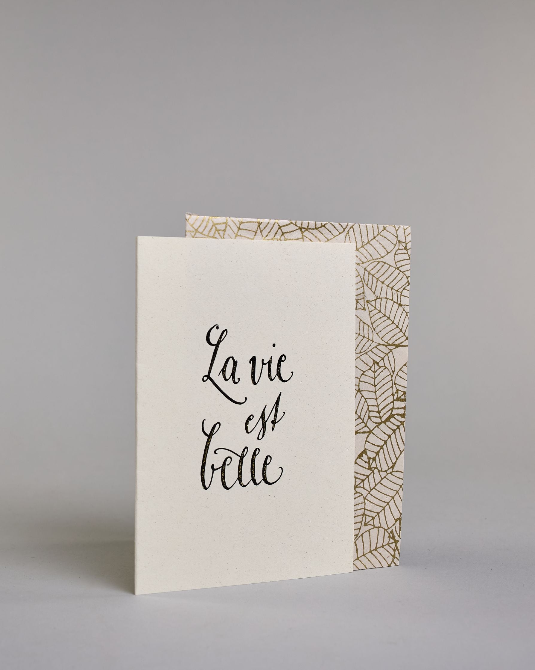Karte auf Französischmit der Aufschrift „La vie est belle“ (übersetzt „Das Leben ist schön“) in schwarzer Handlettering-Schrift auf hellem Hintergrund. Die Kalligarfieaufschrift ist auf drei Zeilen aufgeteilt und in geschwungener Schrift gestaltet. Dazu ein weiß-goldenes handgeschöpftes Kuvert. #3