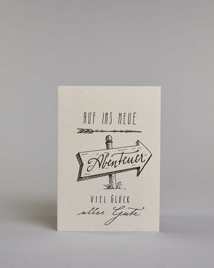 Karte mit der Aufschrift „Auf ins neue Abenteuer – Viel Glück, alles Gute“ in schwarzer Handlettering-Schrift. In der Mitte ist ein handgezeichnetes Schild in der Form eines Pfeiles der nach rechts weist, auf diesem befindet sich das Wort "Abenteuer", "AUF INS NEUE" steht darüber, "VIEL GLÜCK" und "alles Gute" darunter auf hellem Hintergrund. Dazu ein grün-goldenes handgeschöpftes Kuvert. #1