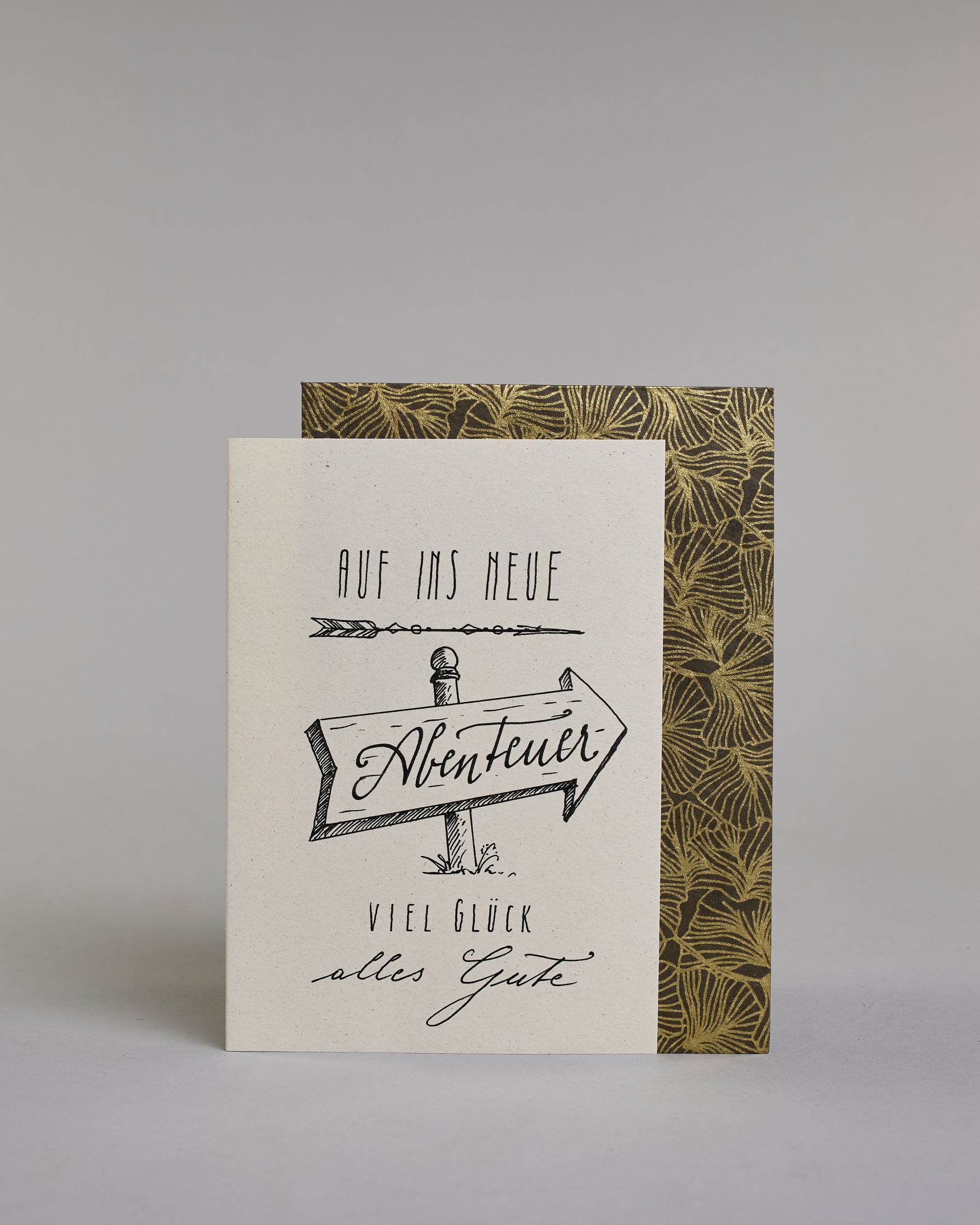 Karte mit der Aufschrift „Auf ins neue Abenteuer – Viel Glück, alles Gute“ in schwarzer Handlettering-Schrift. In der Mitte ist ein handgezeichnetes Schild in der Form eines Pfeiles der nach rechts weist, auf diesem befindet sich das Wort "Abenteuer", "AUF INS NEUE" steht darüber, "VIEL GLÜCK" und "alles Gute" darunter auf hellem Hintergrund. Dazu ein grün-goldenes handgeschöpftes Kuvert. #2