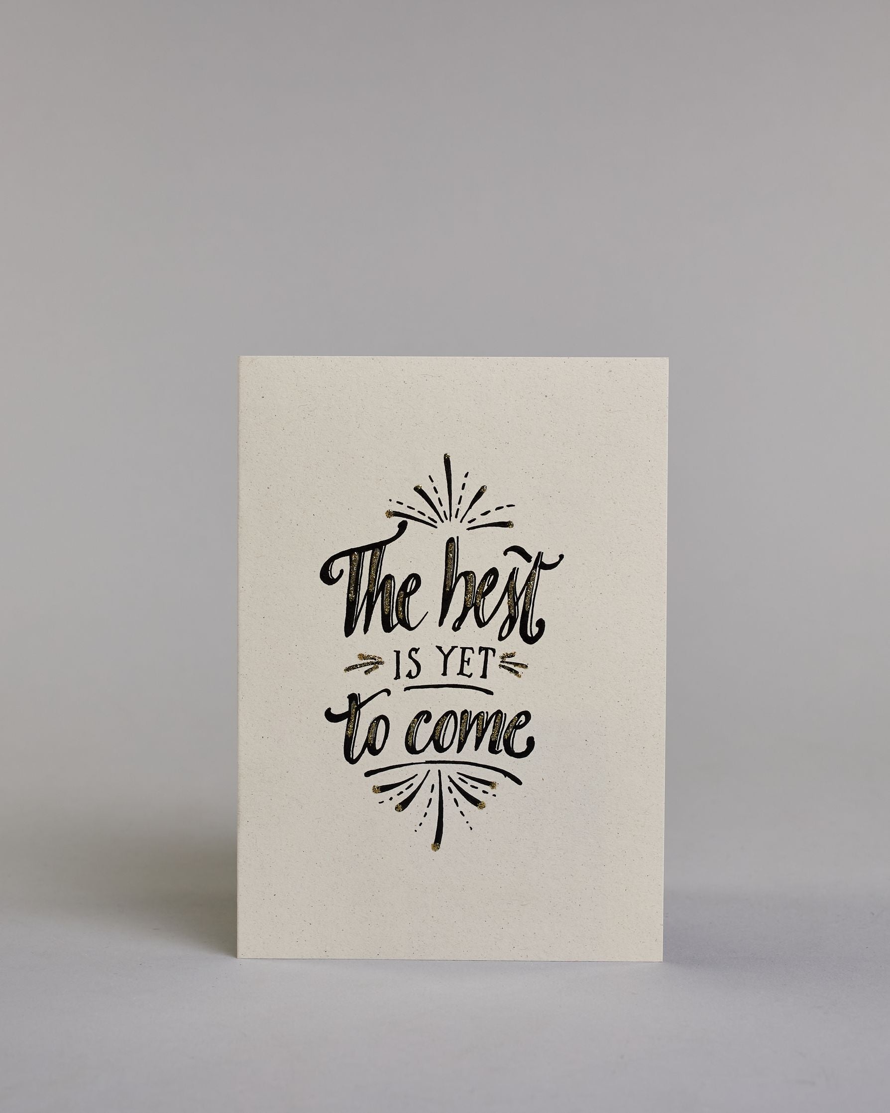 Karte in Englisch mit der Aufschrift „The best is yet to come“ in schwarzer Handlettering-Schrift, teilweise mit goldfarbenen Akzenten hervorgehoben. Über und unter dem Text sind kleine stilisierte, ebenfalls verglitzerte Feuerwerksmotive abgebildet. Dazu ein weiß-goldenes handgeschöpftes Kuvert. #1