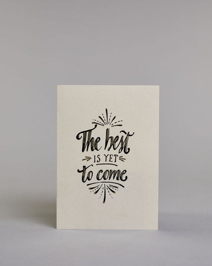 Karte in Englisch mit der Aufschrift „The best is yet to come“ in schwarzer Handlettering-Schrift, teilweise mit goldfarbenen Akzenten hervorgehoben. Über und unter dem Text sind kleine stilisierte, ebenfalls verglitzerte Feuerwerksmotive abgebildet. Dazu ein weiß-goldenes handgeschöpftes Kuvert. #1