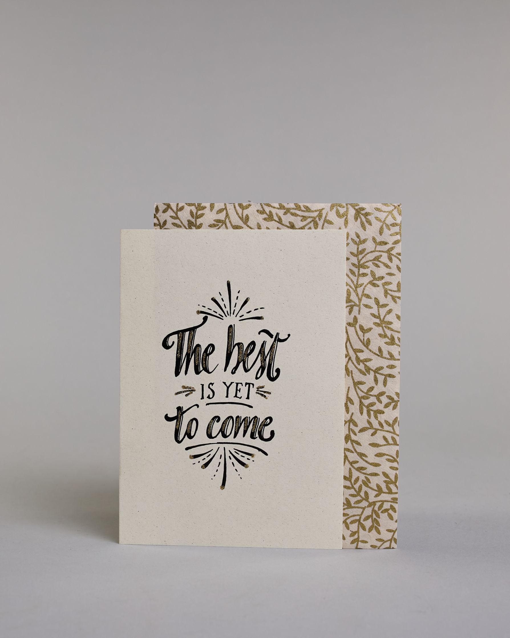 Karte in Englisch mit der Aufschrift „The best is yet to come“ in schwarzer Handlettering-Schrift, teilweise mit goldfarbenen Akzenten hervorgehoben. Über und unter dem Text sind kleine stilisierte, ebenfalls verglitzerte Feuerwerksmotive abgebildet. Dazu ein weiß-goldenes handgeschöpftes Kuvert. #2