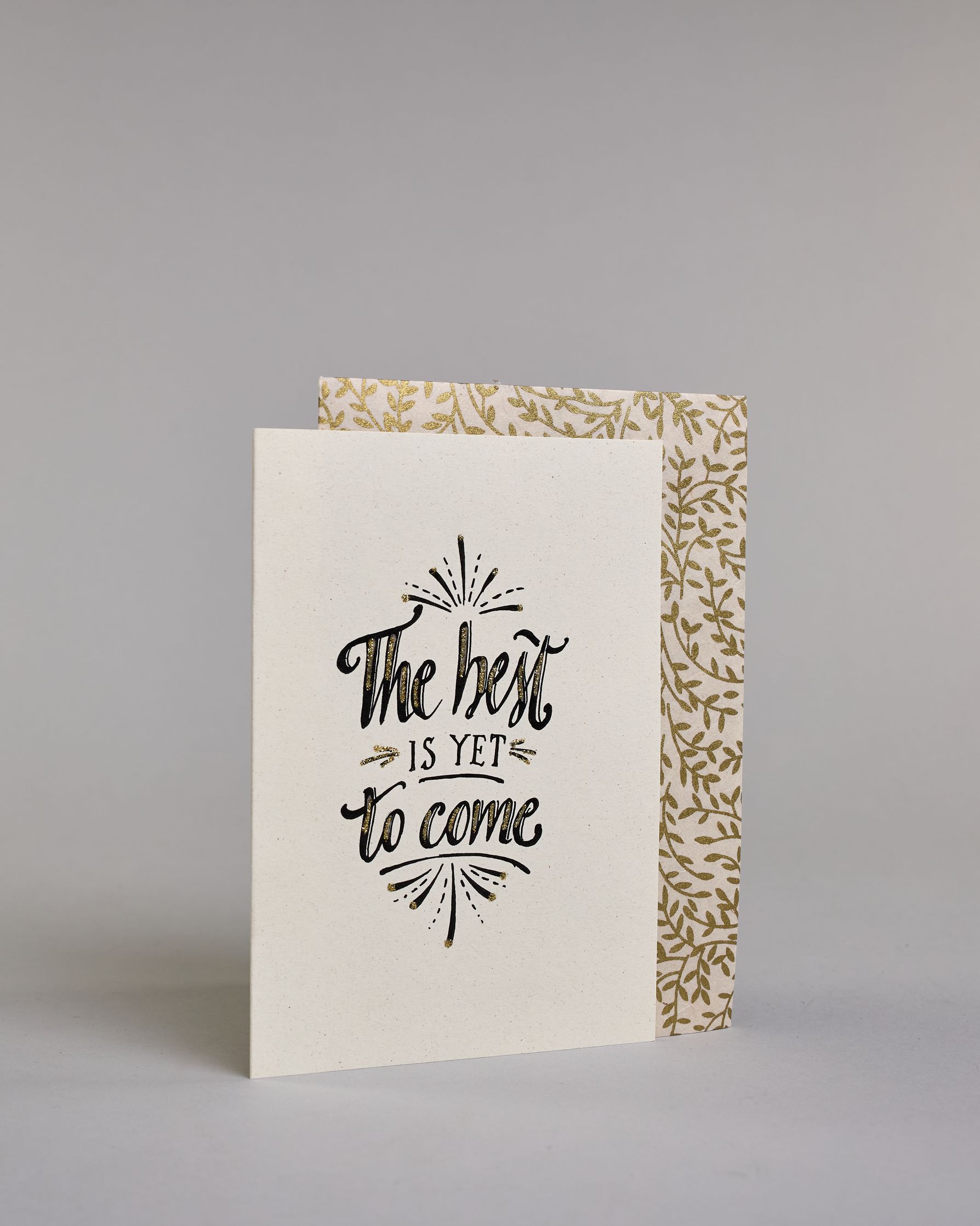 Karte in Englisch mit der Aufschrift „The best is yet to come“ in schwarzer Handlettering-Schrift, teilweise mit goldfarbenen Akzenten hervorgehoben. Über und unter dem Text sind kleine stilisierte, ebenfalls verglitzerte Feuerwerksmotive abgebildet. Dazu ein weiß-goldenes handgeschöpftes Kuvert. #3