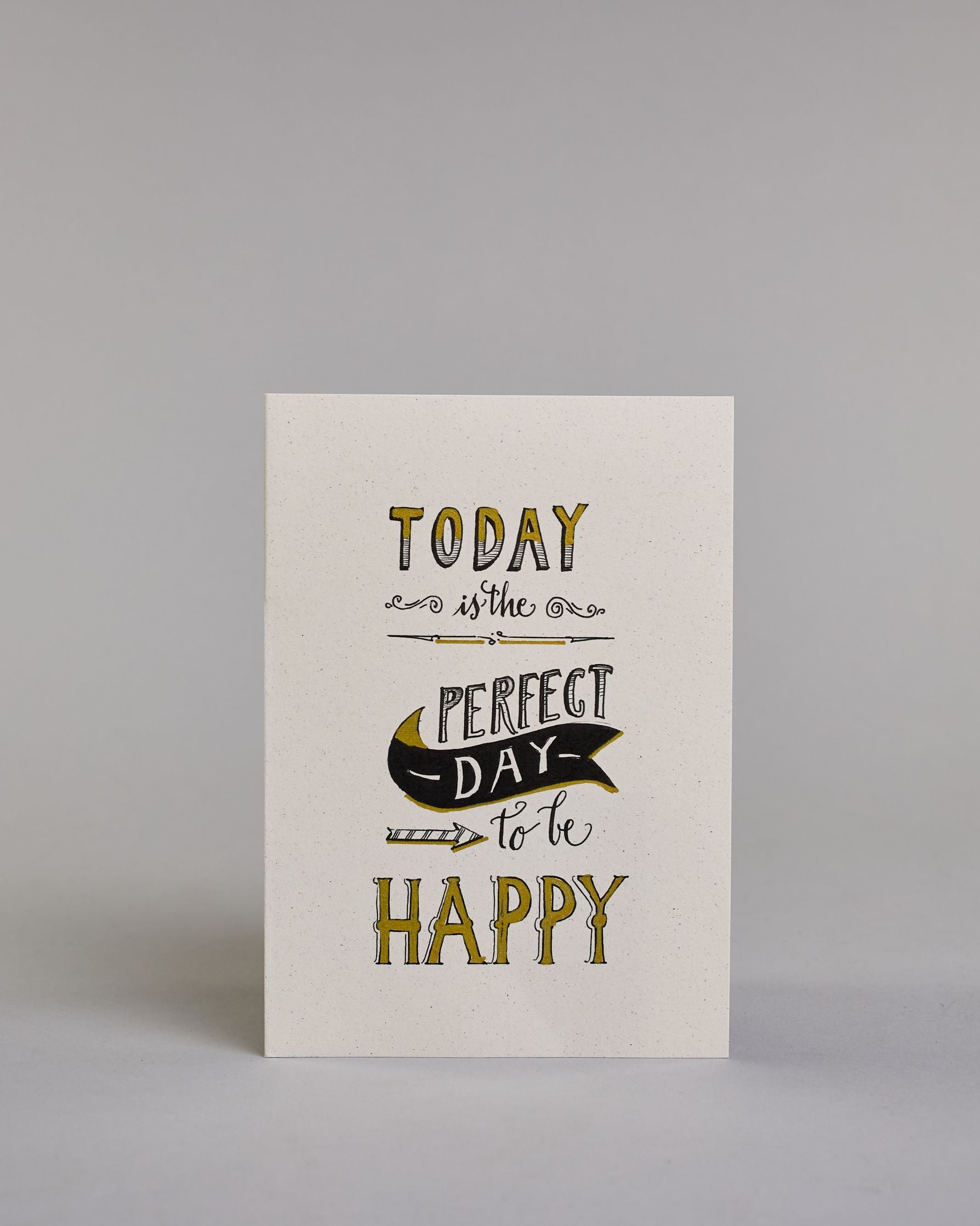 Karte in Englisch mit der Aufschrift „Today is the perfect day to be happy“ in dekorativer Handlettering-Schrift. Die Wörter „TODAY“, „PERFECT DAY“ und „HAPPY“ sind mit goldfarbenen Akzenten hervorgehoben. Ein schwarzes Bannerl ergänzet das Design, in diesem befindet sich das Wort "DAY", darunter ist ein nach rechts, zu den Wörtern "to be" zeigender Pfeil auf hellem Hintergrund. Dazu ein grün-goldenes handgeschöpftes Kuvert. #1