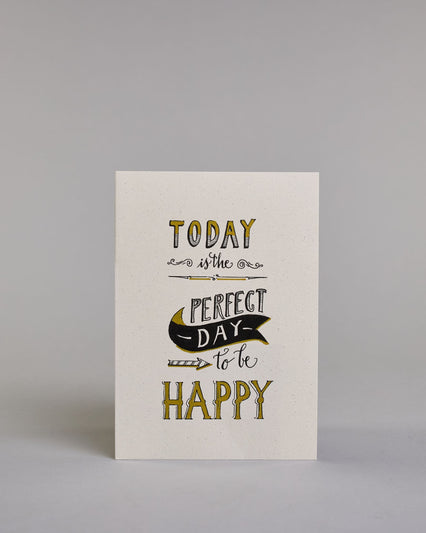 Karte in Englisch mit der Aufschrift „Today is the perfect day to be happy“ in dekorativer Handlettering-Schrift. Die Wörter „TODAY“, „PERFECT DAY“ und „HAPPY“ sind mit goldfarbenen Akzenten hervorgehoben. Ein schwarzes Bannerl ergänzet das Design, in diesem befindet sich das Wort "DAY", darunter ist ein nach rechts, zu den Wörtern "to be" zeigender Pfeil auf hellem Hintergrund. Dazu ein grün-goldenes handgeschöpftes Kuvert. #1