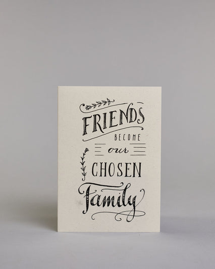 Karte in Englisch mit der Aufschrift „Friends become our chosen family“ in schwarzer Handlettering-Schrift auf hellem Hintergrund. Kleine dekorative Elemente wie Linien, Zweige und Schnörkel verzieren den Text. Dazu ein grün-goldenes handgeschöpftes Kuvert. #1