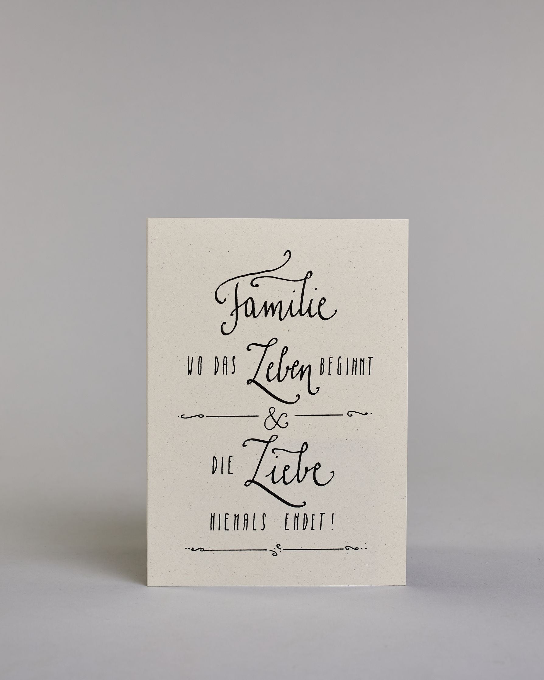 Karte mit der Aufschrift „Familie wo das Leben beginnt & die Liebe niemals endet!“ in schwarzer Handlettering-Schrift auf hellem Hintergrund. Feine Linienornamente rahmen den Text dekorativ ein. Das Design ist schlicht und elegant. Herausgehoben sind die Wörter "Familie", "Leben" und "Liebe". Dazu ein grün-goldenes handgeschöpftes Kuvert. #1