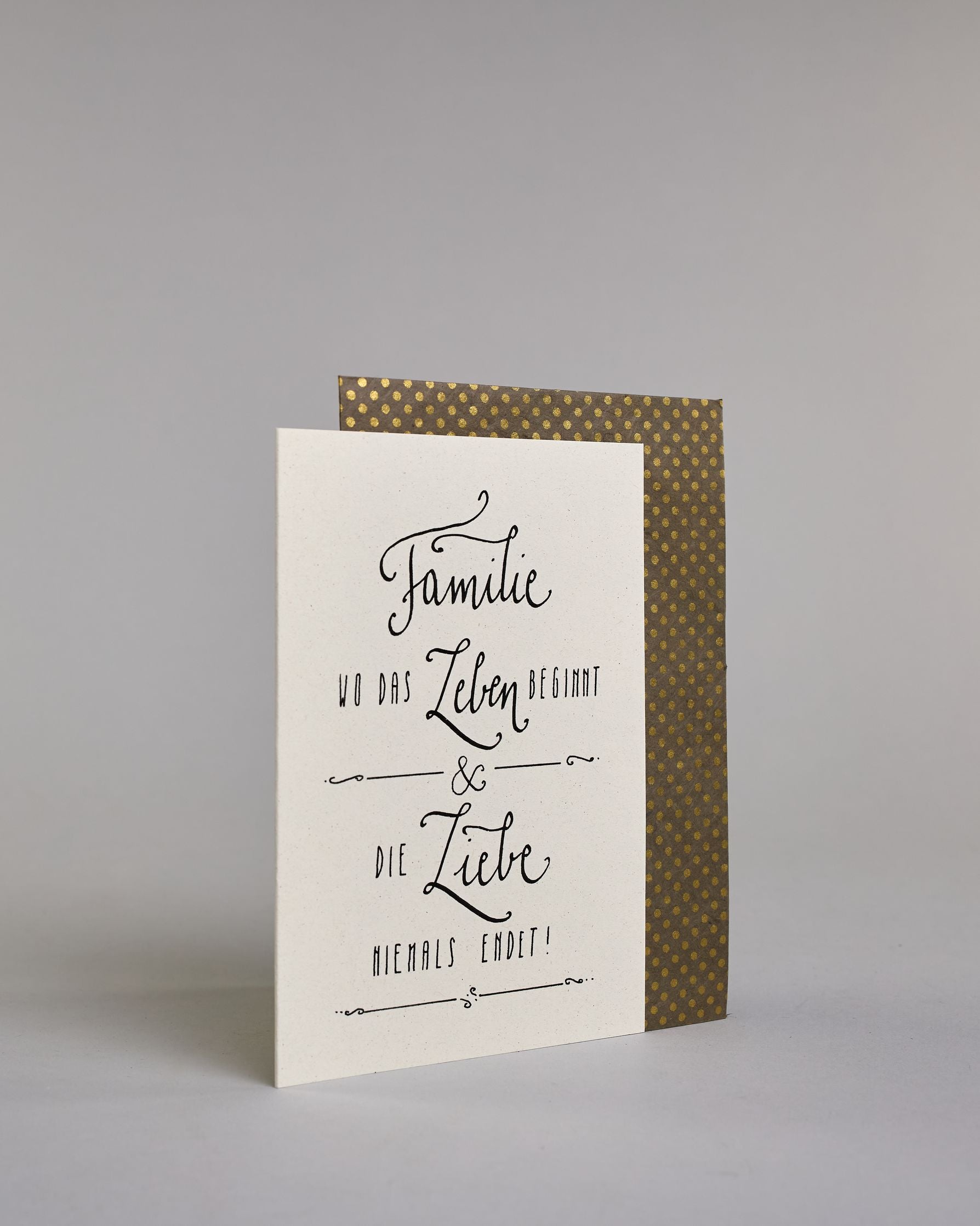 Karte mit der Aufschrift „Familie wo das Leben beginnt & die Liebe niemals endet!“ in schwarzer Handlettering-Schrift auf hellem Hintergrund. Feine Linienornamente rahmen den Text dekorativ ein. Das Design ist schlicht und elegant. Herausgehoben sind die Wörter "Familie", "Leben" und "Liebe". Dazu ein grün-goldenes handgeschöpftes Kuvert. #3