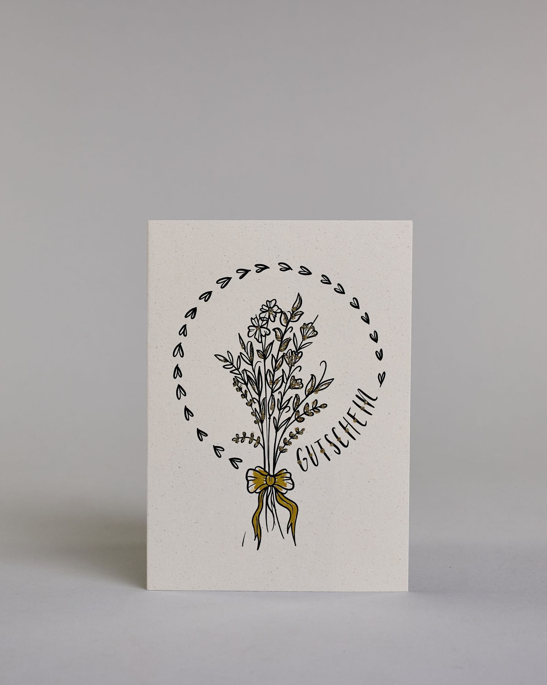 Gutschein-Karte mit einem gezeichneten Blumenstrauß aus filigranen Blüten und Zweigen, zusammengebunden mit einer goldfarbenen Schleife. Um den Strauß verläuft ein Herzkranz. Rechts neben dem Strauß steht das Wort „Gutschein“ in schwarzer Schrift in den Kranz eingearbeitet auf hellem Hintergrund. Dazu ein grün-goldenes handgeschöpftes Kuvert. #1