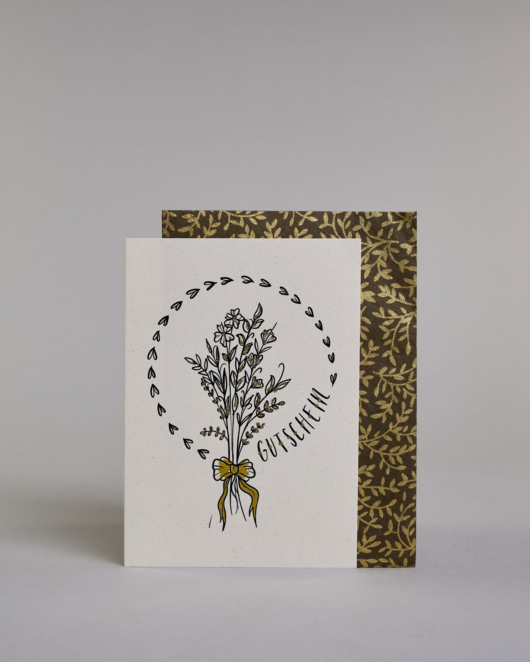 Gutschein-Karte mit einem gezeichneten Blumenstrauß aus filigranen Blüten und Zweigen, zusammengebunden mit einer goldfarbenen Schleife. Um den Strauß verläuft ein Herzkranz. Rechts neben dem Strauß steht das Wort „Gutschein“ in schwarzer Schrift in den Kranz eingearbeitet auf hellem Hintergrund. Dazu ein grün-goldenes handgeschöpftes Kuvert. #2