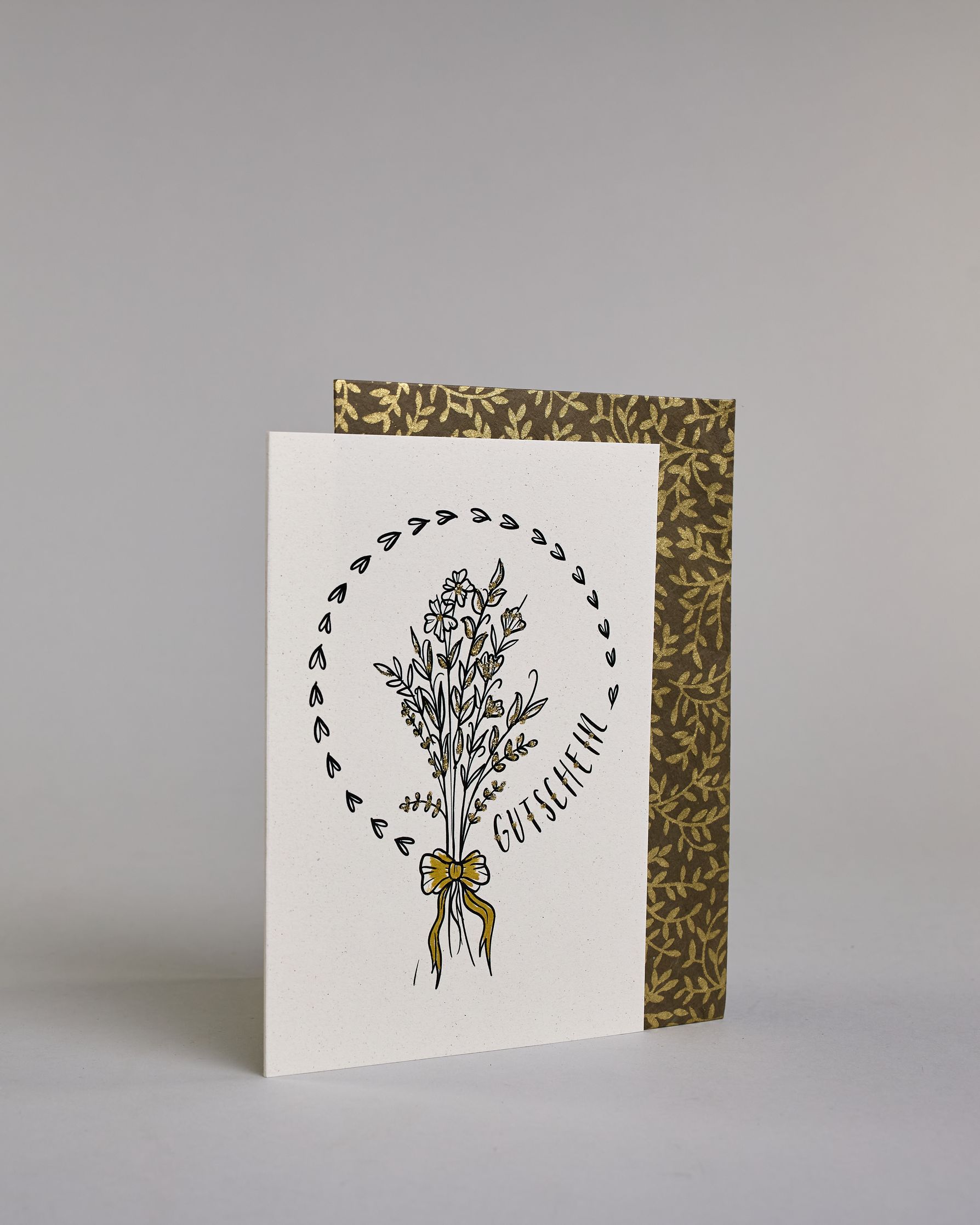 Gutschein-Karte mit einem gezeichneten Blumenstrauß aus filigranen Blüten und Zweigen, zusammengebunden mit einer goldfarbenen Schleife. Um den Strauß verläuft ein Herzkranz. Rechts neben dem Strauß steht das Wort „Gutschein“ in schwarzer Schrift in den Kranz eingearbeitet auf hellem Hintergrund. Dazu ein grün-goldenes handgeschöpftes Kuvert. #3