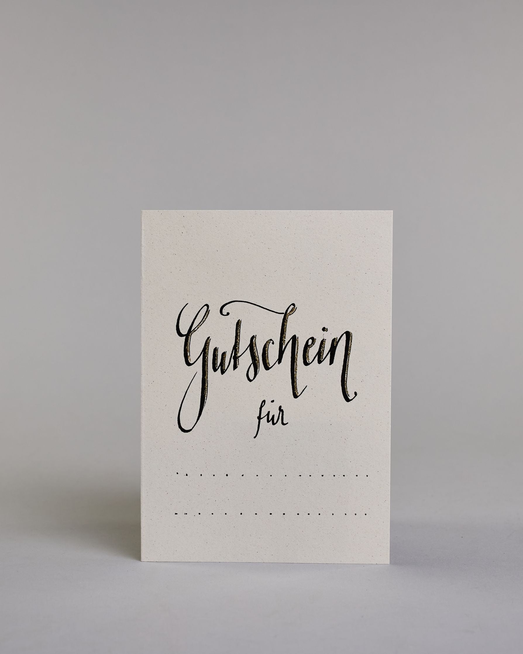Gutschein-Karte mit der Aufschrift „Gutschein für …“ in schwarzer Handlettering-Schrift. Darunter befinden sich gepunktete Linien zum individuellen Ausfüllen auf hellem Hintergrund. Dazu ein grün-goldenes handgeschöpftes Kuvert. #1