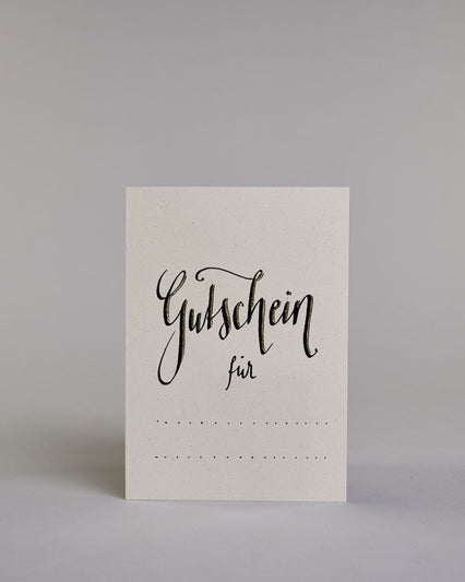 Gutschein-Karte mit der Aufschrift „Gutschein für …“ in schwarzer Handlettering-Schrift. Darunter befinden sich gepunktete Linien zum individuellen Ausfüllen auf hellem Hintergrund. Dazu ein grün-goldenes handgeschöpftes Kuvert. #1