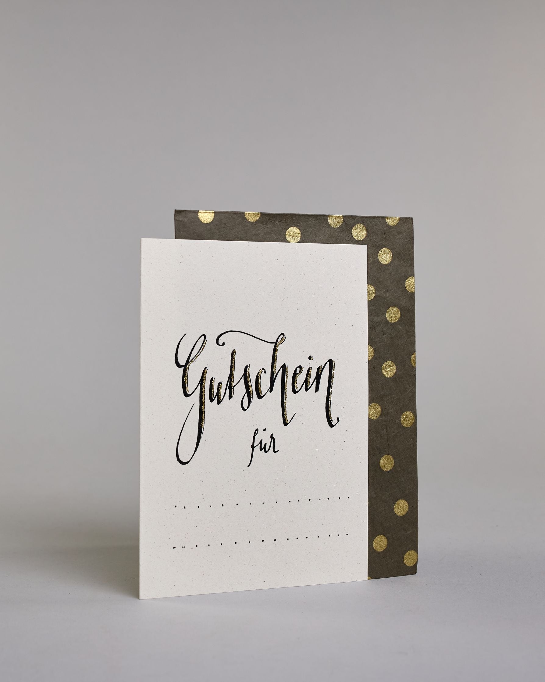 Gutschein-Karte mit der Aufschrift „Gutschein für …“ in schwarzer Handlettering-Schrift. Darunter befinden sich gepunktete Linien zum individuellen Ausfüllen auf hellem Hintergrund. Dazu ein grün-goldenes handgeschöpftes Kuvert. #3
