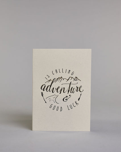Karte mit der Aufschrift „Adventure is calling & good luck“ in schwarzer Handlettering-Schrift auf hellem Hintergrund. Über dem Wort „adventure“, welches mittig auf der Karte zu finden ist sind stilisierte Berge gezeichnet, darunter ein kleiner Weg. Zwei Pfeile rahmen das Design seitlich ein, was zu einer kreisrunden Optik führt. Dazu ein grün-goldenes handgeschöpftes Kuvert. #1