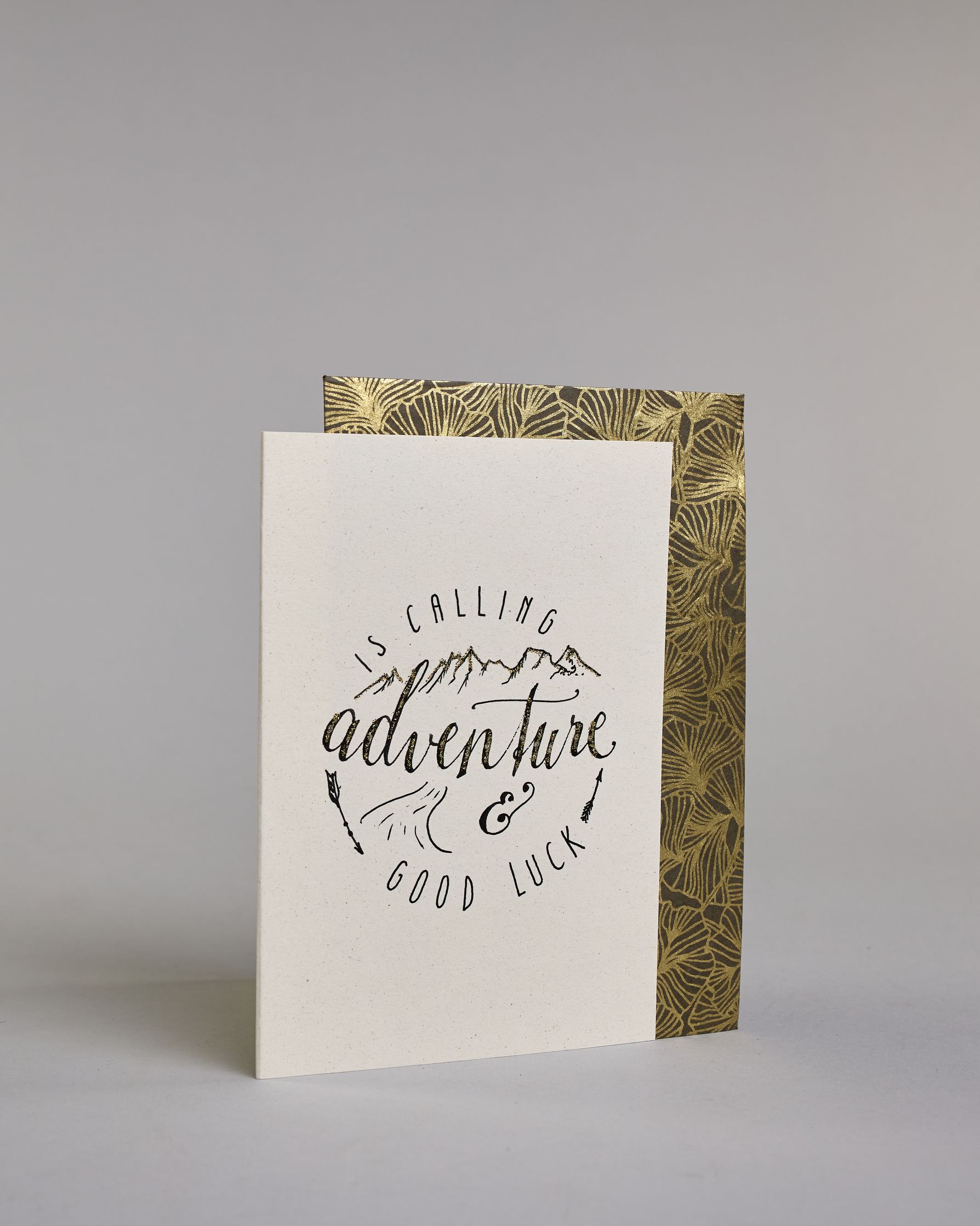 Karte mit der Aufschrift „Adventure is calling & good luck“ in schwarzer Handlettering-Schrift auf hellem Hintergrund. Über dem Wort „adventure“, welches mittig auf der Karte zu finden ist sind stilisierte Berge gezeichnet, darunter ein kleiner Weg. Zwei Pfeile rahmen das Design seitlich ein, was zu einer kreisrunden Optik führt. Dazu ein grün-goldenes handgeschöpftes Kuvert. #3