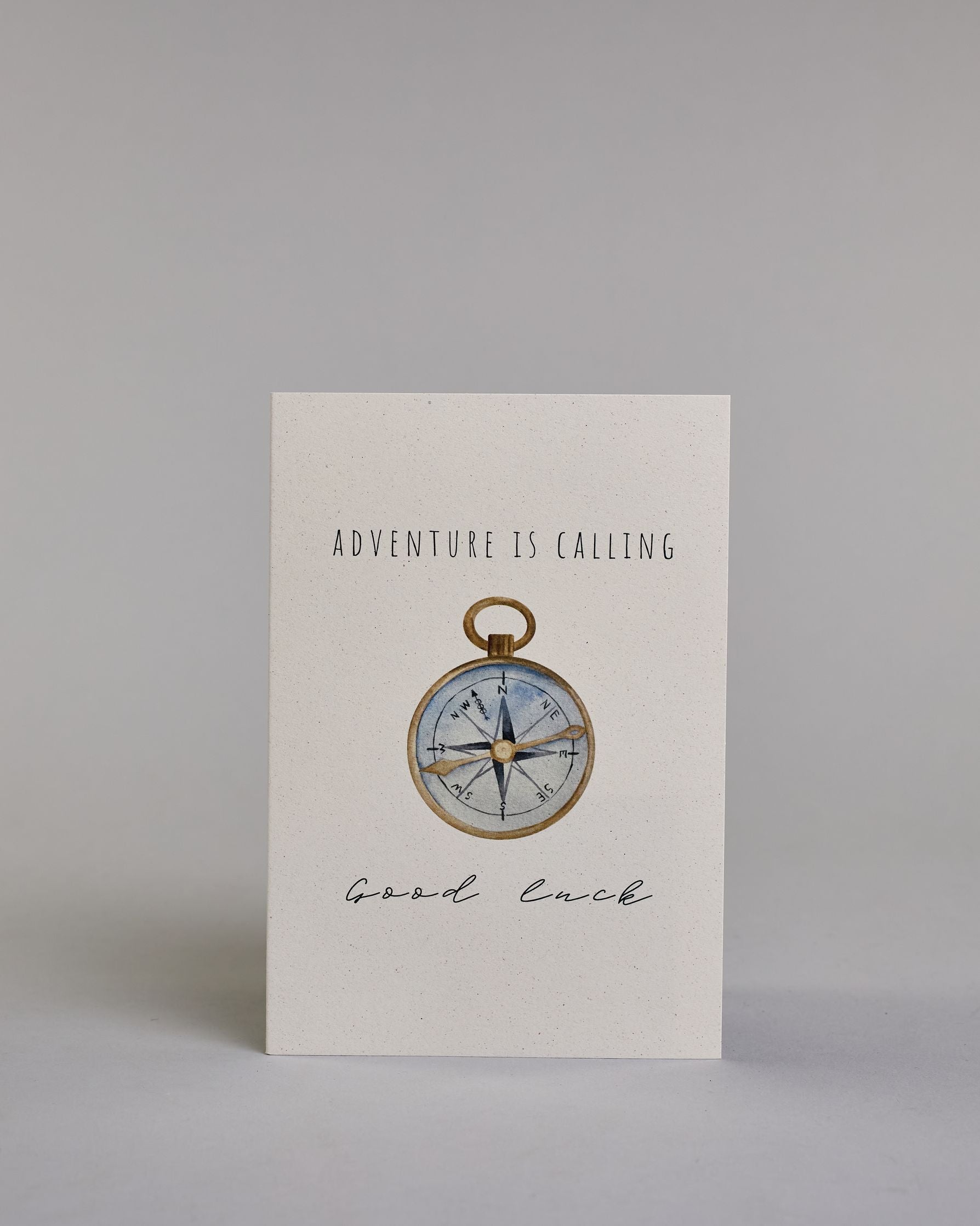Karte mit der Aufschrift „Adventure is calling Good luck“ in schwarzer Kalligrafieaufschrift. In der Mitte ist eine detailreich gezeichnete Kompass-Illustration mit goldenem Rand und goldener Nadel abgebildet, die Innenseite des Kompasses ist hellblau und die Karte selbst bildet einen hellen Hintergrund. Dazu ein grün-goldenes handgeschöpftes Kuvert. #1
