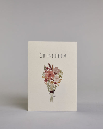 Gutschein-Karte mit der schlichten Aufschrift „Gutschein“ in schwarzer Schrift auf hellem Hintergrund. Darunter ist ein stilisierter Blumenstrauß in gedeckten Rosa-, Rot- und Grüntönen abgebildet, zusammengebunden mit einer schwarzen Schleife.Dazu ein grün-goldenes handgeschöpftes Kuvert. #1