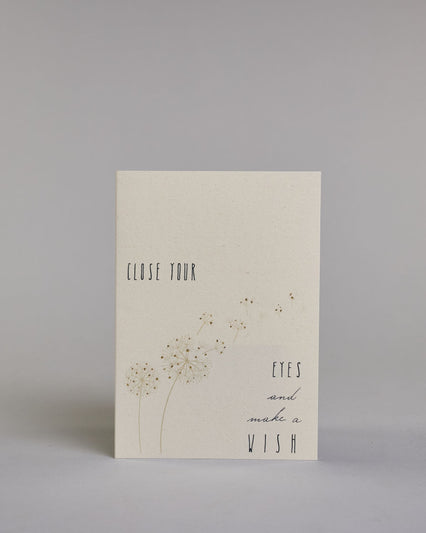 Karte auf Englisch mit der Aufschrift „Close your eyes and make a wish“ in feiner schwarzer Kalligrafieaufschrift auf hellem Hintergrund. Links unten sind zarte Pusteblumen abgebildet, deren Samen sanft quer über die Karte verwehen. Dazu ein weißes handgeschöpftes Kuvert. #1