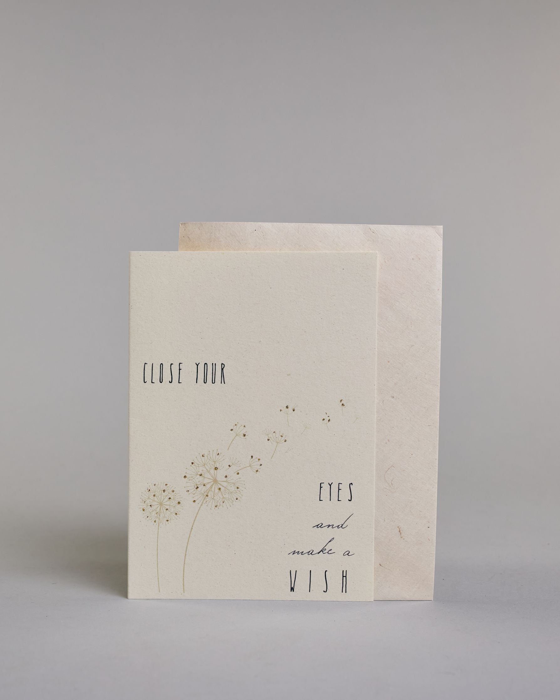 Karte auf Englisch mit der Aufschrift „Close your eyes and make a wish“ in feiner schwarzer Kalligrafieaufschrift auf hellem Hintergrund. Links unten sind zarte Pusteblumen abgebildet, deren Samen sanft quer über die Karte verwehen. Dazu ein weißes handgeschöpftes Kuvert. #2