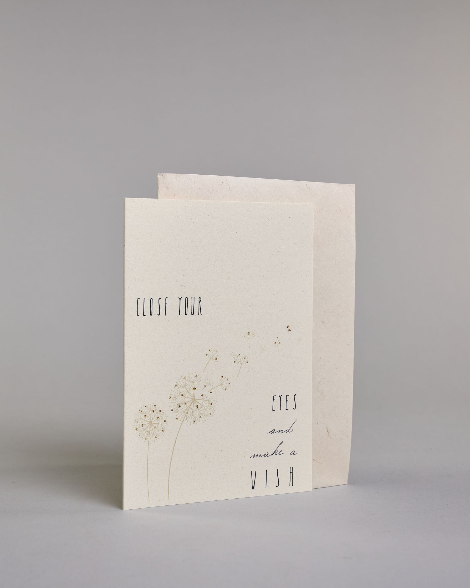Karte auf Englisch mit der Aufschrift „Close your eyes and make a wish“ in feiner schwarzer Kalligrafieaufschrift auf hellem Hintergrund. Links unten sind zarte Pusteblumen abgebildet, deren Samen sanft quer über die Karte verwehen. Dazu ein weißes handgeschöpftes Kuvert. #3