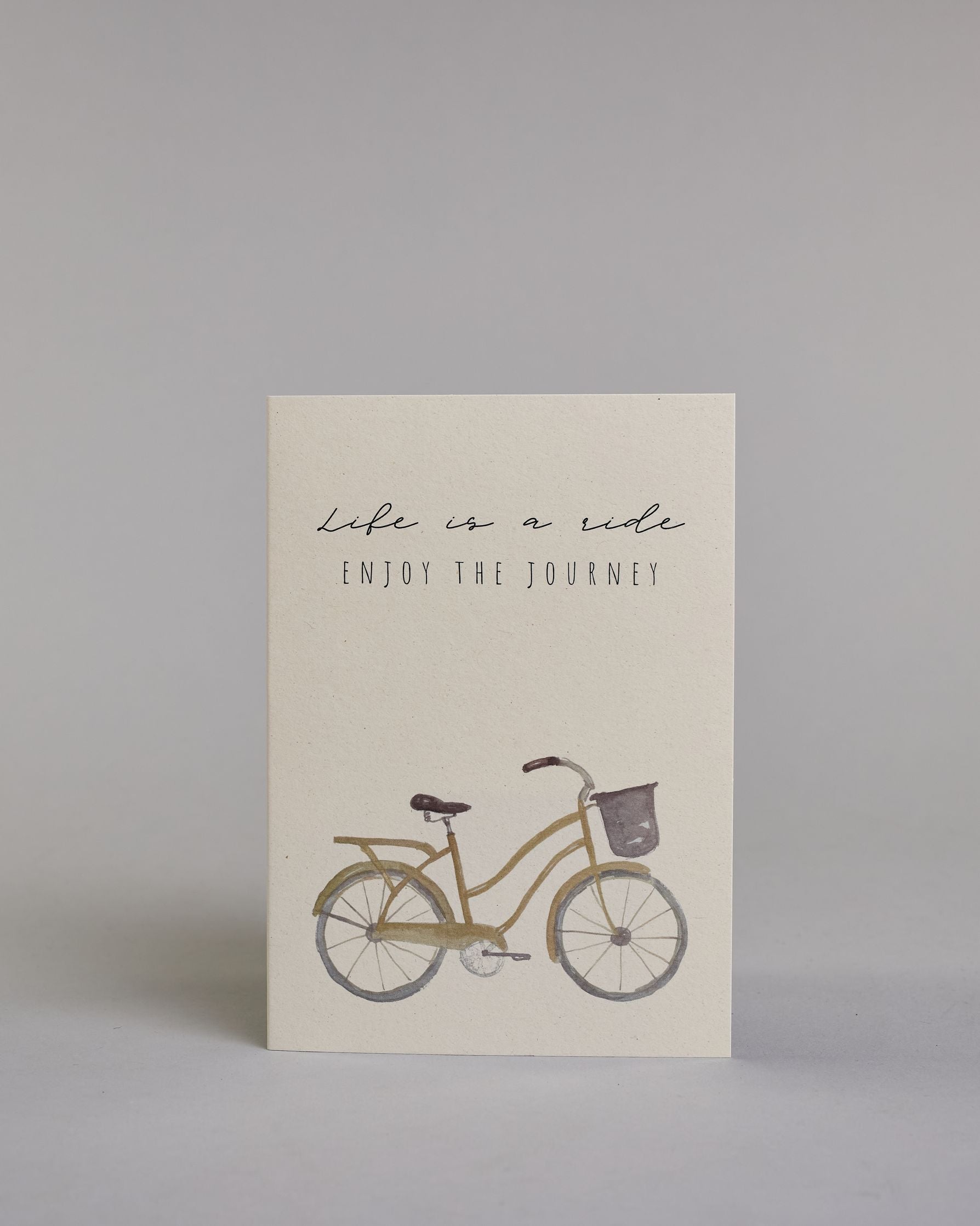 Karte in Englisch mit der Aufschrift „Life is a ride Enjoy the journey“ in feiner schwarzer Schrift auf hellem Hintergrund. Darunter ist ein schlicht illustriertes Fahrrad mit Korb in gedeckten Braun- und Grautönen abgebildet. Dazu ein weißes handgeschöpftes Kuvert. #1