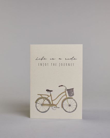 Karte in Englisch mit der Aufschrift „Life is a ride Enjoy the journey“ in feiner schwarzer Schrift auf hellem Hintergrund. Darunter ist ein schlicht illustriertes Fahrrad mit Korb in gedeckten Braun- und Grautönen abgebildet. Dazu ein weißes handgeschöpftes Kuvert. #1