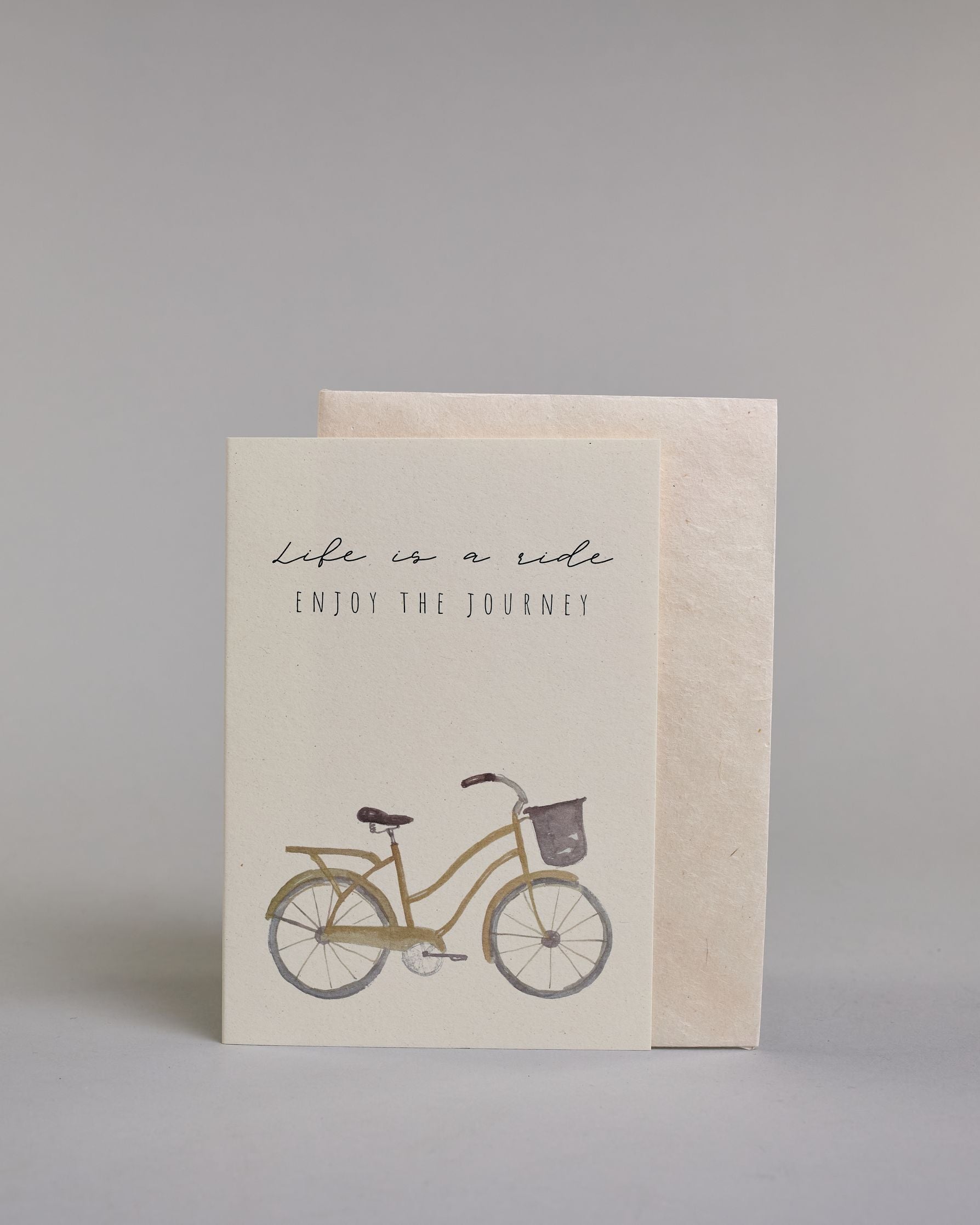 Karte in Englisch mit der Aufschrift „Life is a ride Enjoy the journey“ in feiner schwarzer Schrift auf hellem Hintergrund. Darunter ist ein schlicht illustriertes Fahrrad mit Korb in gedeckten Braun- und Grautönen abgebildet. Dazu ein weißes handgeschöpftes Kuvert. #2