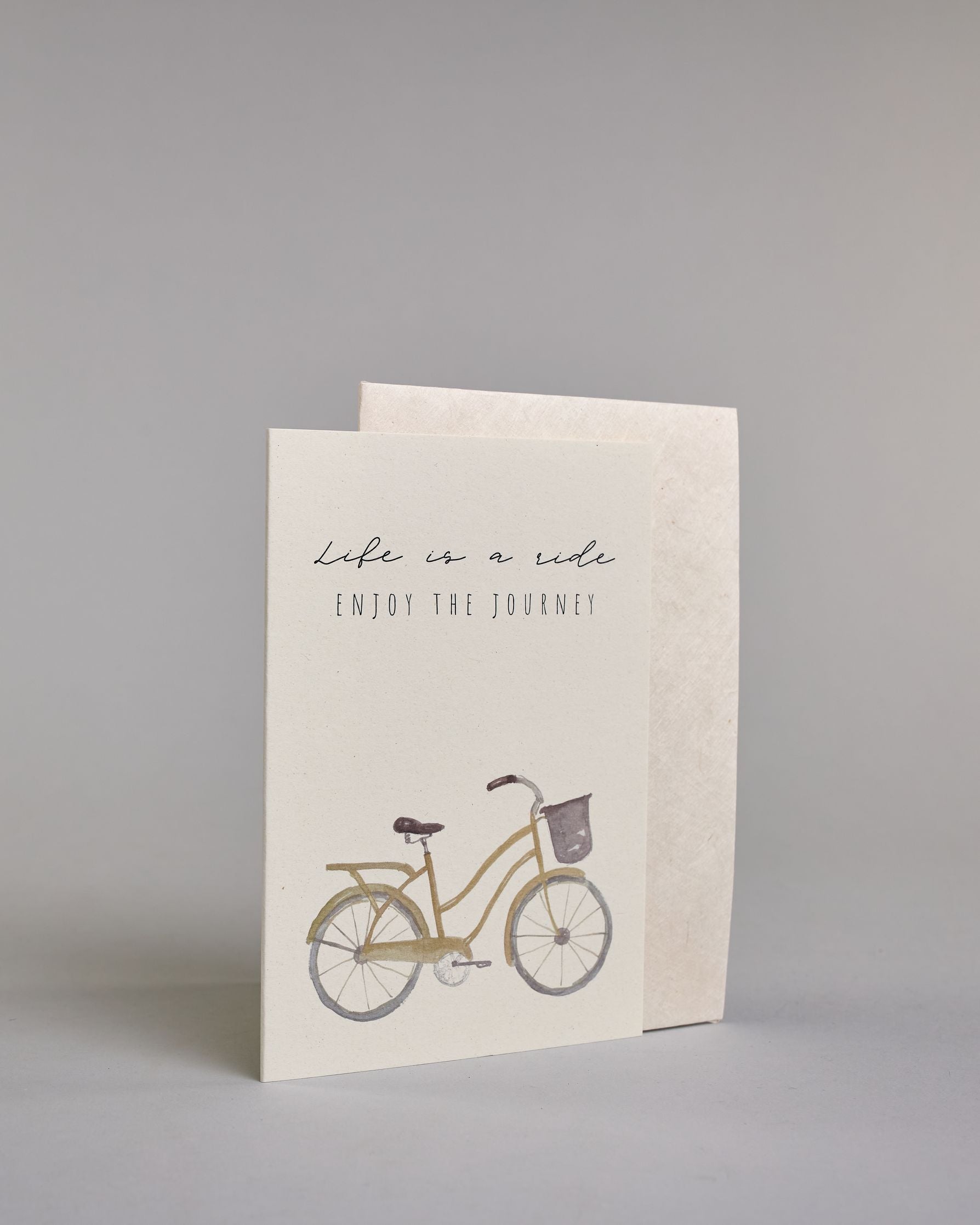 Karte in Englisch mit der Aufschrift „Life is a ride Enjoy the journey“ in feiner schwarzer Schrift auf hellem Hintergrund. Darunter ist ein schlicht illustriertes Fahrrad mit Korb in gedeckten Braun- und Grautönen abgebildet. Dazu ein weißes handgeschöpftes Kuvert. #3