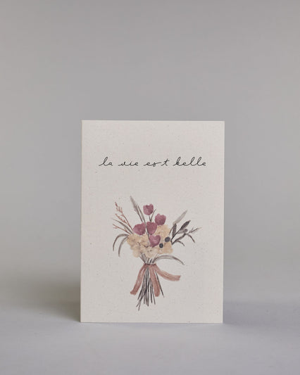 Karte mit der französischen Aufschrift „la vie est belle“ (übersetzt „das Leben ist schön“) in feiner schwarzer Kalligrafieaufschrift. Darunter ist ein zarter Blumenstrauß in gedeckten Beige- und Rosatönen abgebildet, locker zusammengebunden mit einer Schleife. Dazu ein weißes handgeschöpftes Kuvert. #1