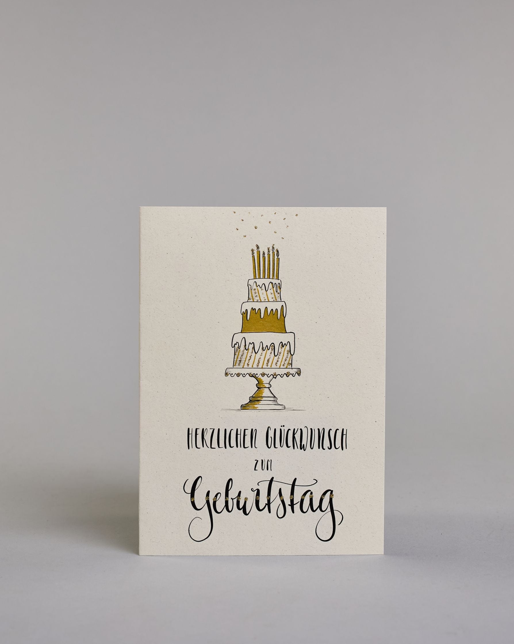 Glückwunschkarte zum Geburtstag mit schwarzer Kalligrafieaufschrift "Herzlichen Glückwunsch zum Geburtstag", darüber eine dreistückige Geburtstagstorte auf einem Tortenständer mit sechs Kerzen auf dem obersten Tortenstock. Torte, Tortenständer, als auch Kerzen sind mit Gold und Golzglitzer verziert, über den Kerzen sind in Goldglitzer Funken angedeutet. Der Text ist in drei Zeilen aufgeteilt und alles ist in einer dickeren geschwungenen Schrift gehalten. Erst "Herzlichen Glückwunsch" eher kleiner gehalten,…