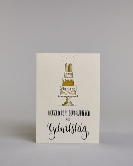 Glückwunschkarte zum Geburtstag mit schwarzer Kalligrafieaufschrift "Herzlichen Glückwunsch zum Geburtstag", darüber eine dreistückige Geburtstagstorte auf einem Tortenständer mit sechs Kerzen auf dem obersten Tortenstock. Torte, Tortenständer, als auch Kerzen sind mit Gold und Golzglitzer verziert, über den Kerzen sind in Goldglitzer Funken angedeutet. Der Text ist in drei Zeilen aufgeteilt und alles ist in einer dickeren geschwungenen Schrift gehalten. Erst "Herzlichen Glückwunsch" eher kleiner gehalten,…