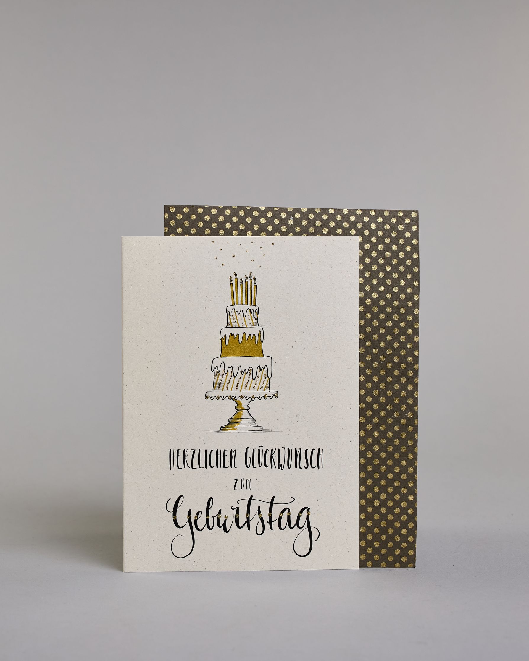 Glückwunschkarte zum Geburtstag mit schwarzer Kalligrafieaufschrift "Herzlichen Glückwunsch zum Geburtstag", darüber eine dreistückige Geburtstagstorte auf einem Tortenständer mit sechs Kerzen auf dem obersten Tortenstock. Torte, Tortenständer, als auch Kerzen sind mit Gold und Golzglitzer verziert, über den Kerzen sind in Goldglitzer Funken angedeutet. Der Text ist in drei Zeilen aufgeteilt und alles ist in einer dickeren geschwungenen Schrift gehalten. Erst "Herzlichen Glückwunsch" eher kleiner gehalten,…