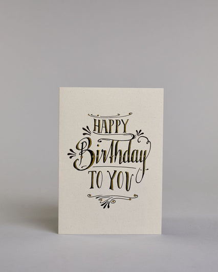 Glückwunschkarte in Englisch zum Geburtstag mit schwarzer Kalligrafieaufschrift "Happy Birthday to you". Der Text ist in drei Zeilen aufgeteilt, welche wiefolge gegliedert sind: "HAPPY", darunter "Birthday", darunter "TO YOU". Die Worte sind mit schwungvollen Linien und Verzierungen umgeben und alles ist mit Gold akzentuiert. Alles ist auf hellem Hintergrund. Dazu ein grün-goldenes handgeschöpftes Kuvert. #1